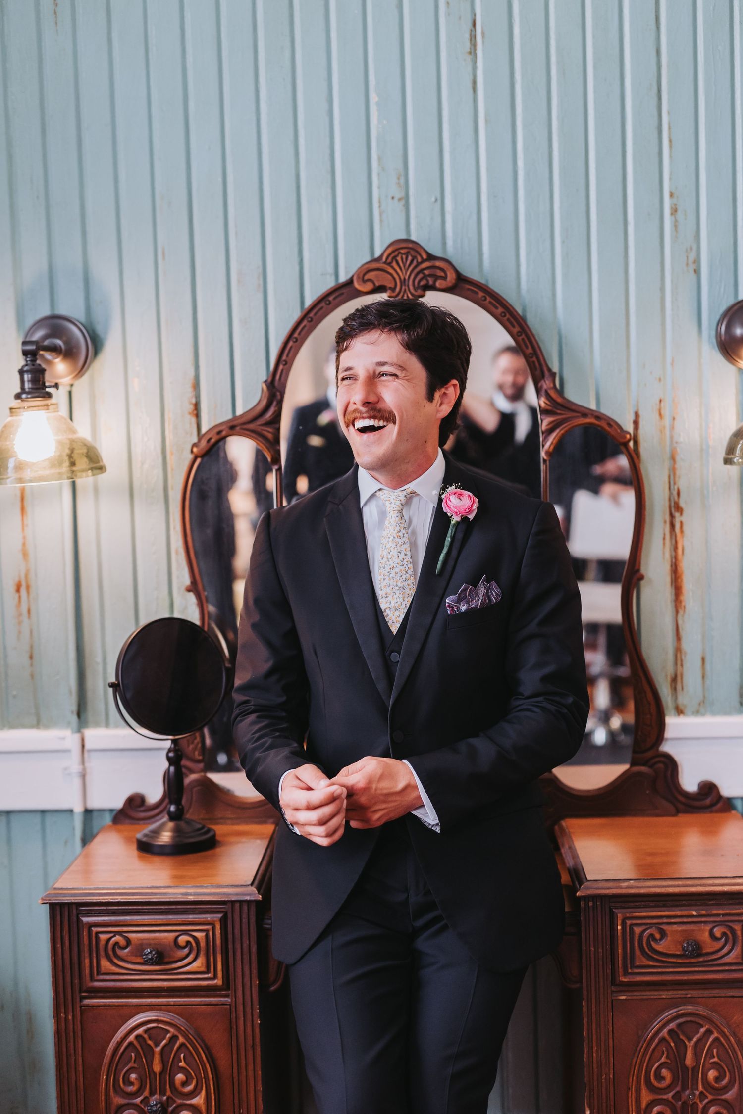 Dapper groom in black three-piece suit with pink boutonniere laughing joyfully before antique wooden dresser.
