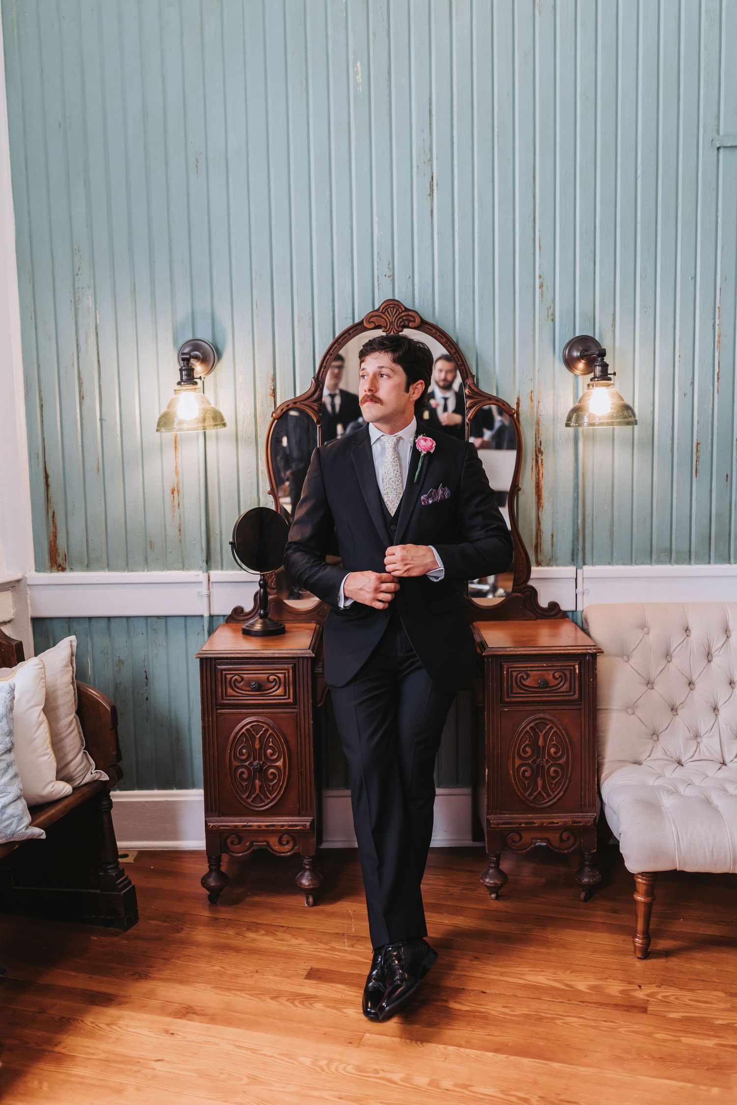 Dapper groom in black suit with pink boutonniere poses by vintage vanity in elegant blue-walled room.