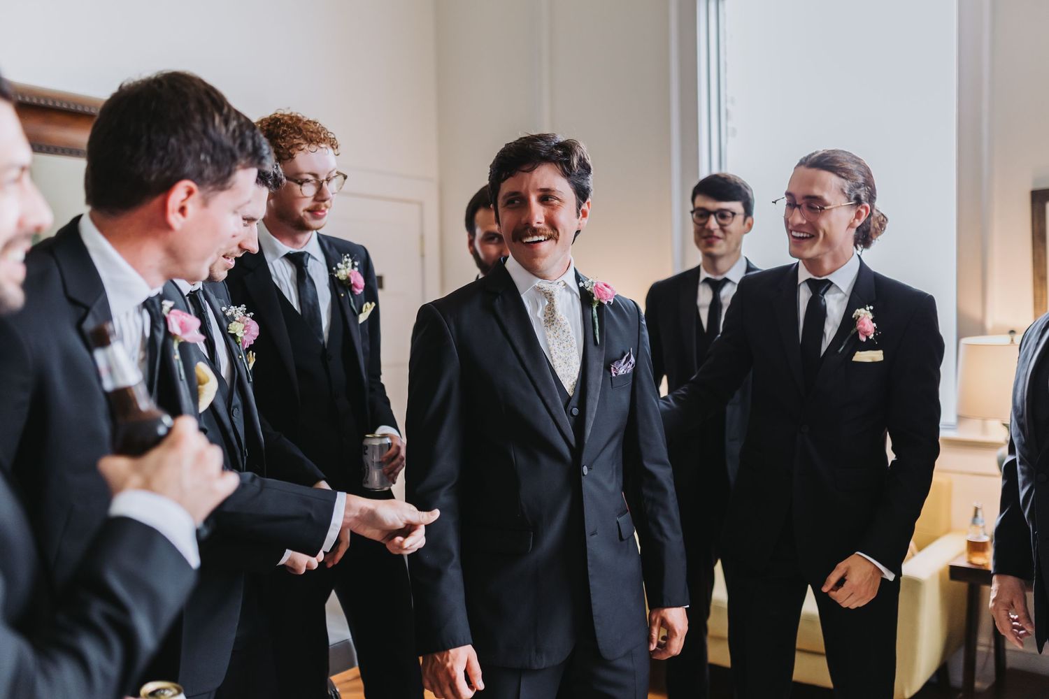 A groom in a black suit laughs with his groomsmen during wedding day preparations indoors.