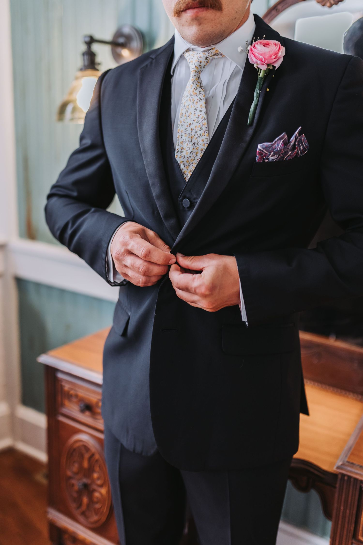 Groom in black tuxedo buttoning jacket with pink boutonniere and floral pocket square.