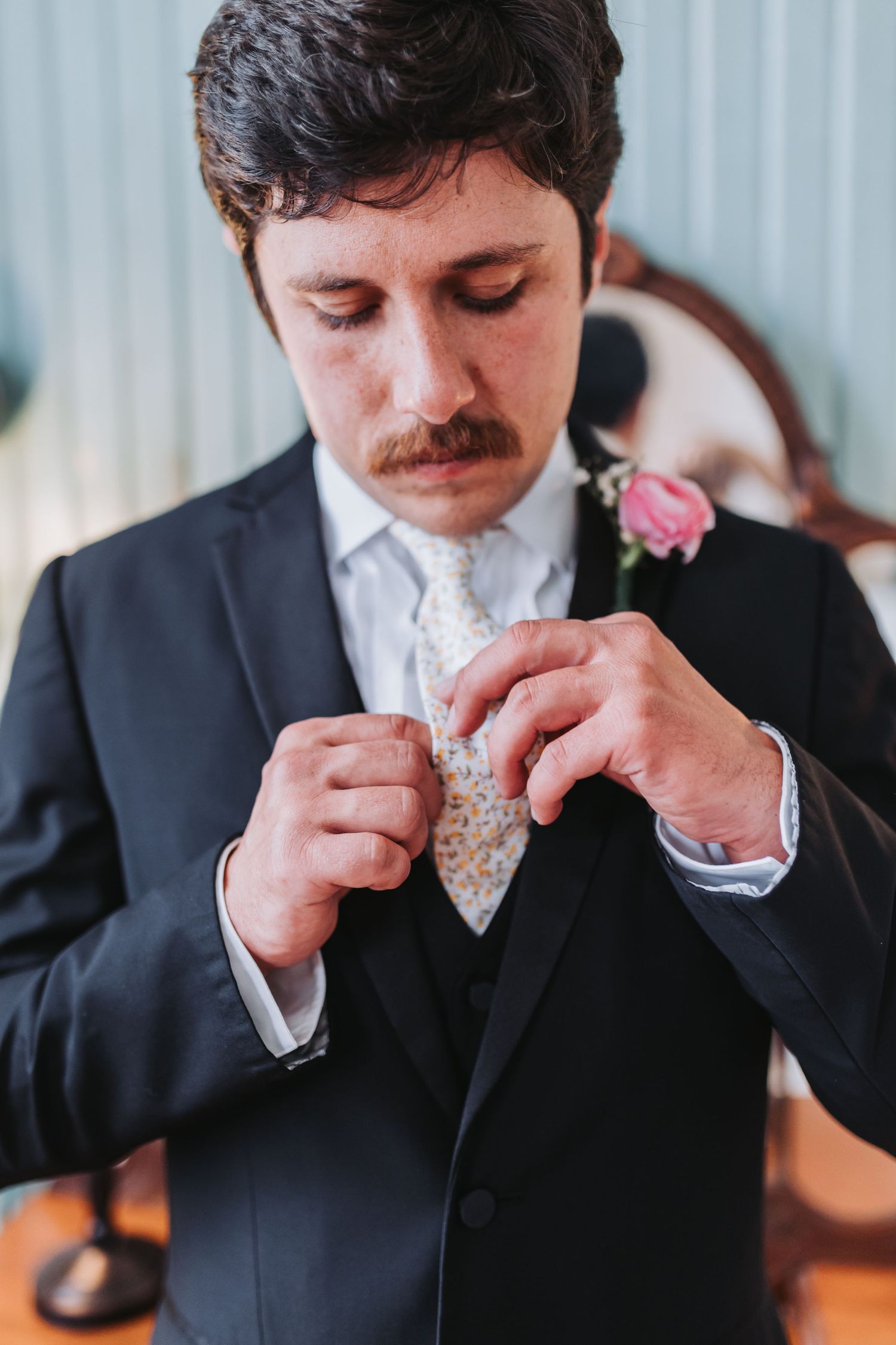 Dapper groom with mustache adjusts his floral tie while getting ready for his wedding day.