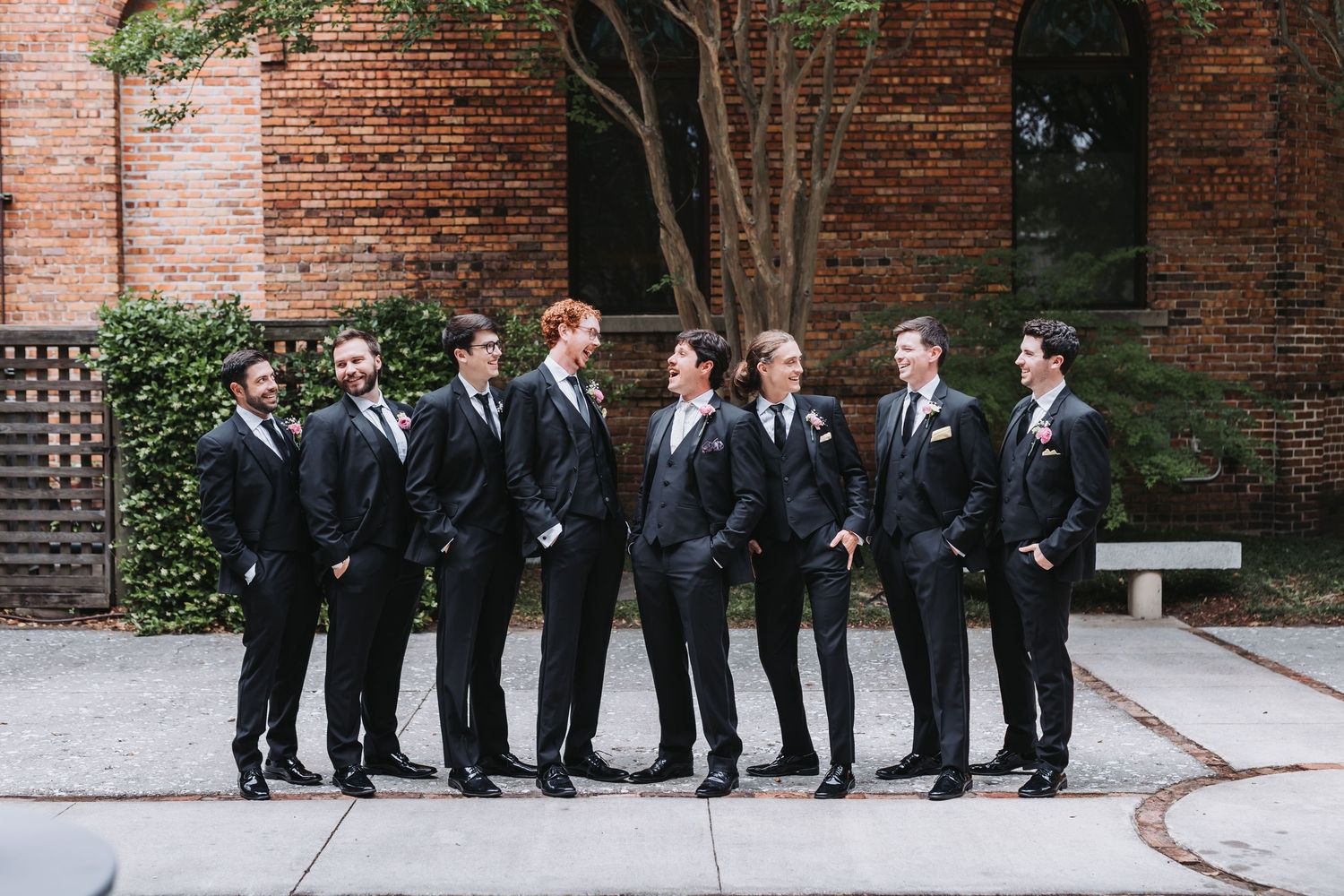 Eight groomsmen in matching black suits pose casually outdoors against a brick building backdrop.