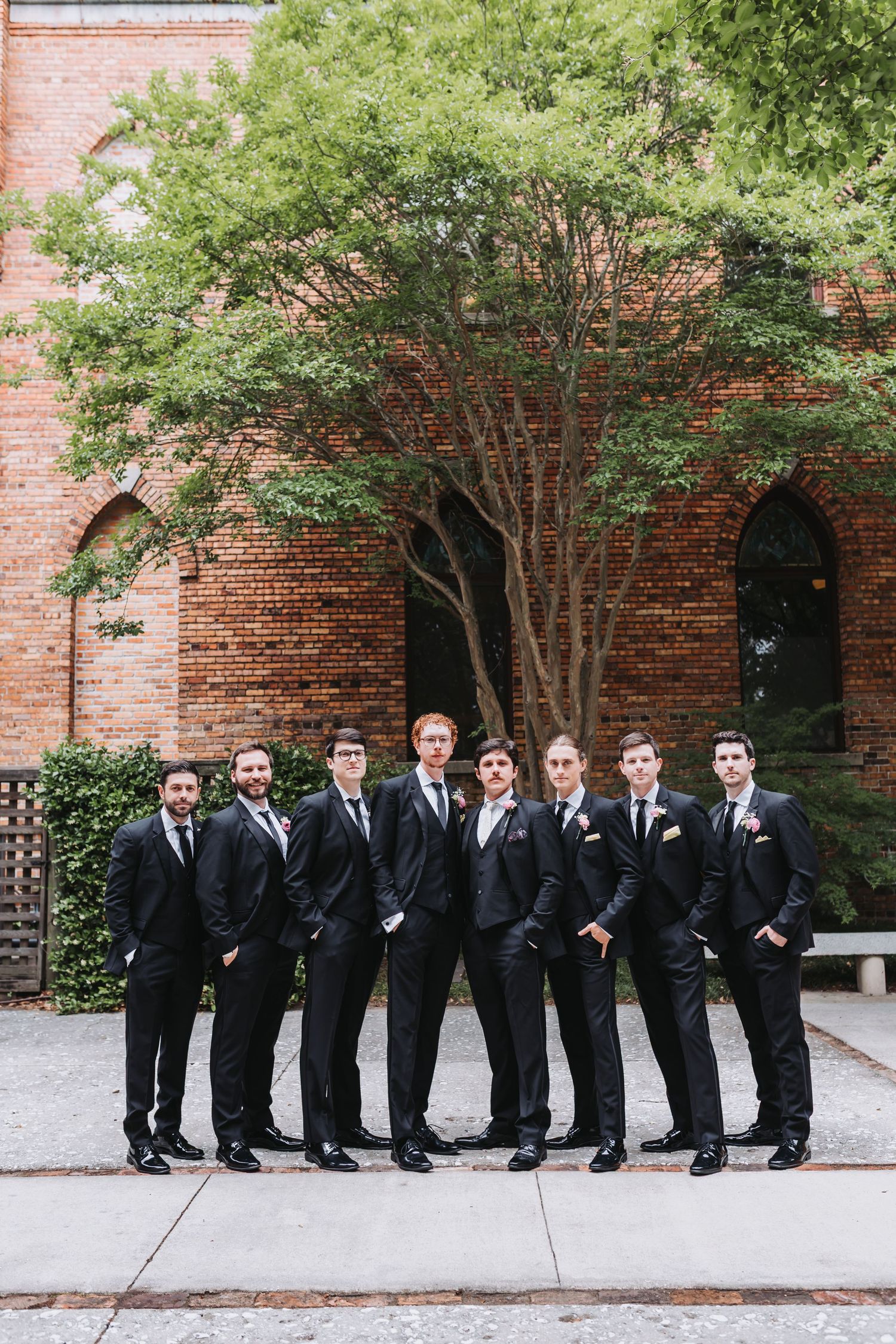Groomsmen in black suits with white pocket squares pose in front of brick church for wedding.