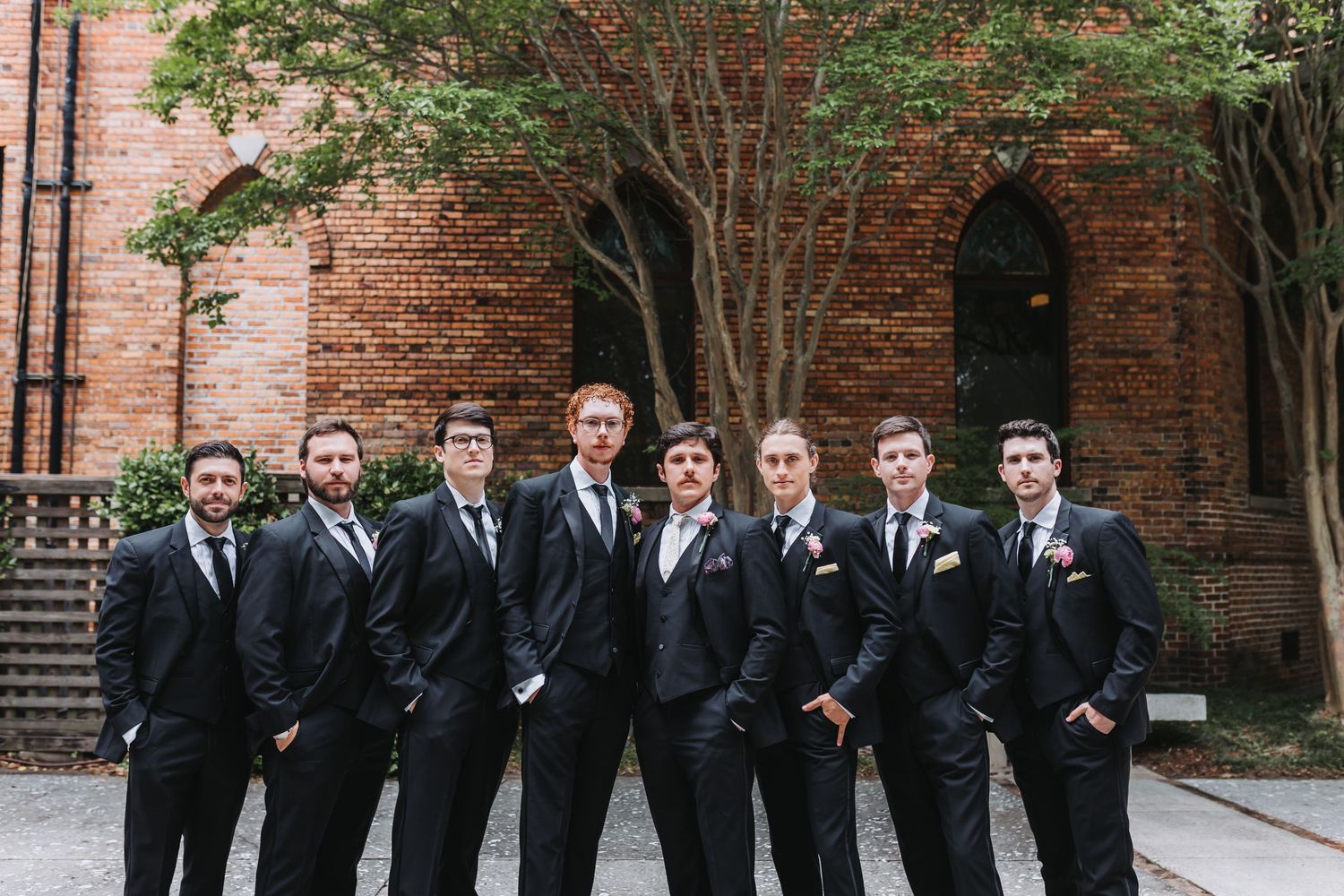 Eight groomsmen in matching black suits pose together outside brick building for wedding party photo.