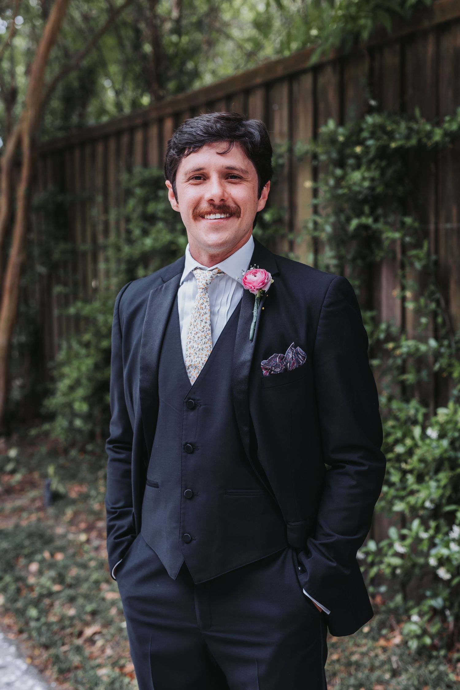 Smiling groom in black three-piece suit with floral tie and pink boutonniere posing outdoors near wooden fence.