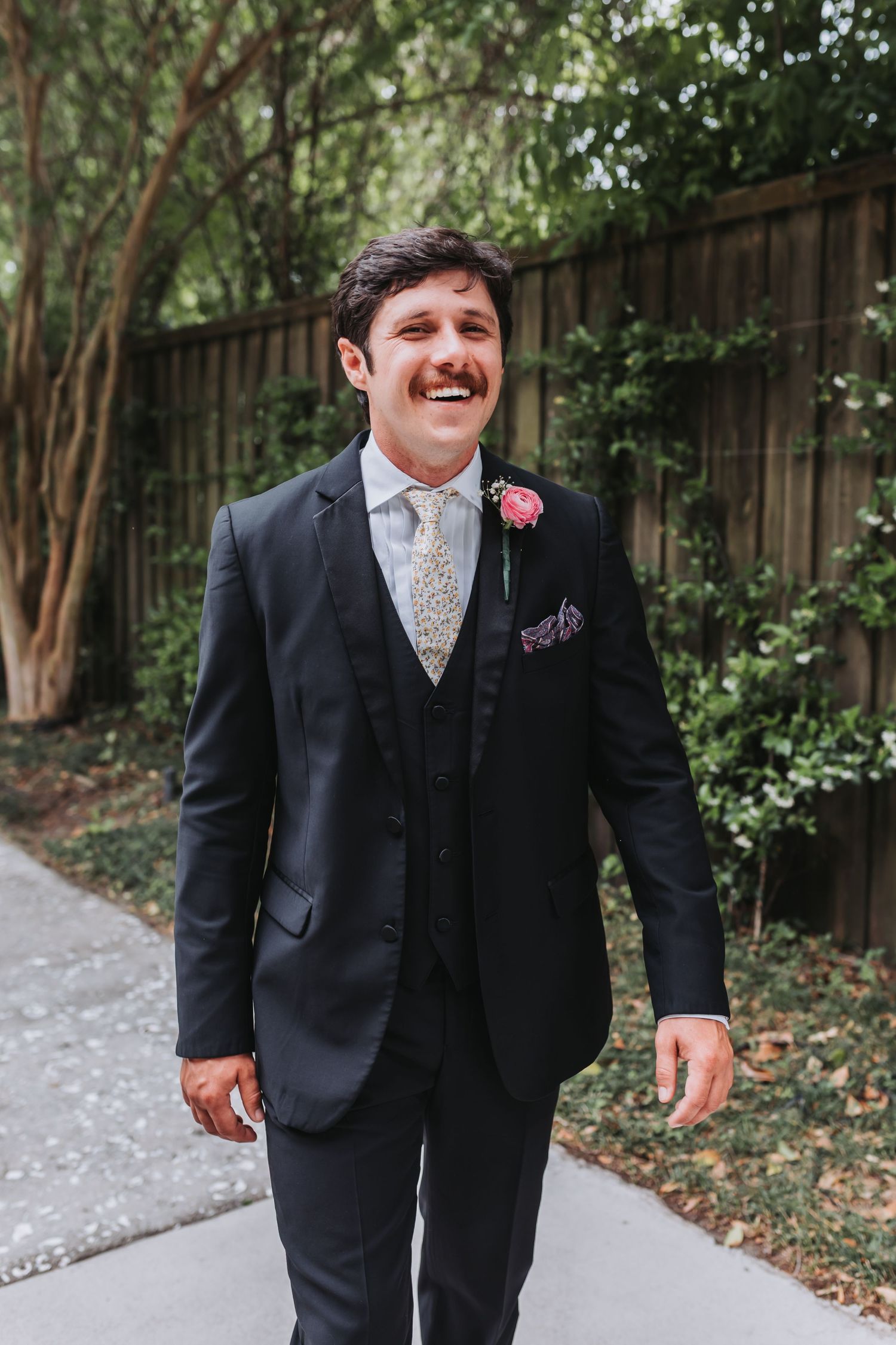 Smiling groom in black three-piece suit with floral boutonniere walks outdoors on wedding day.