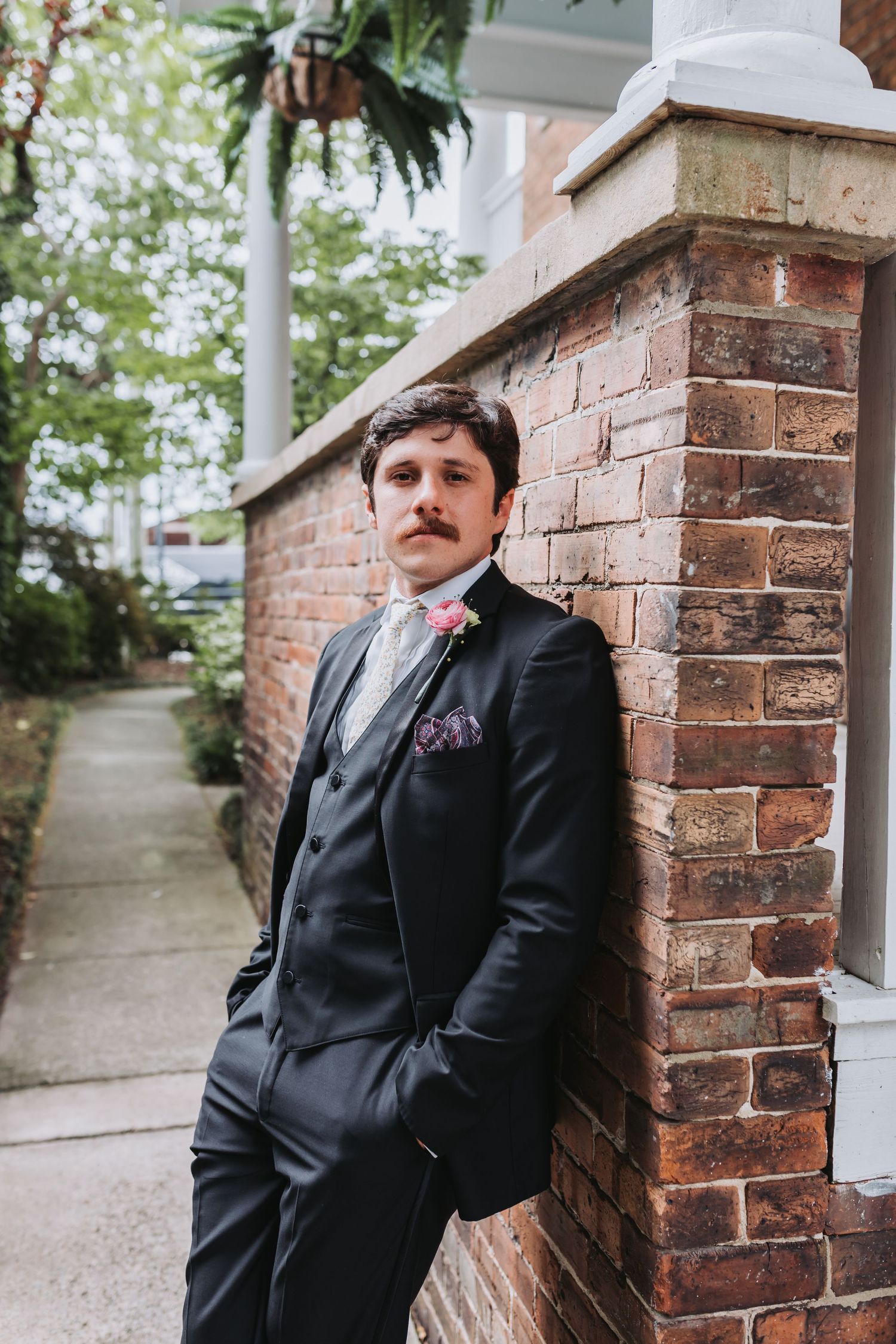 Stylish groom with mustache leans against a brick wall wearing a black suit with pink boutonniere.