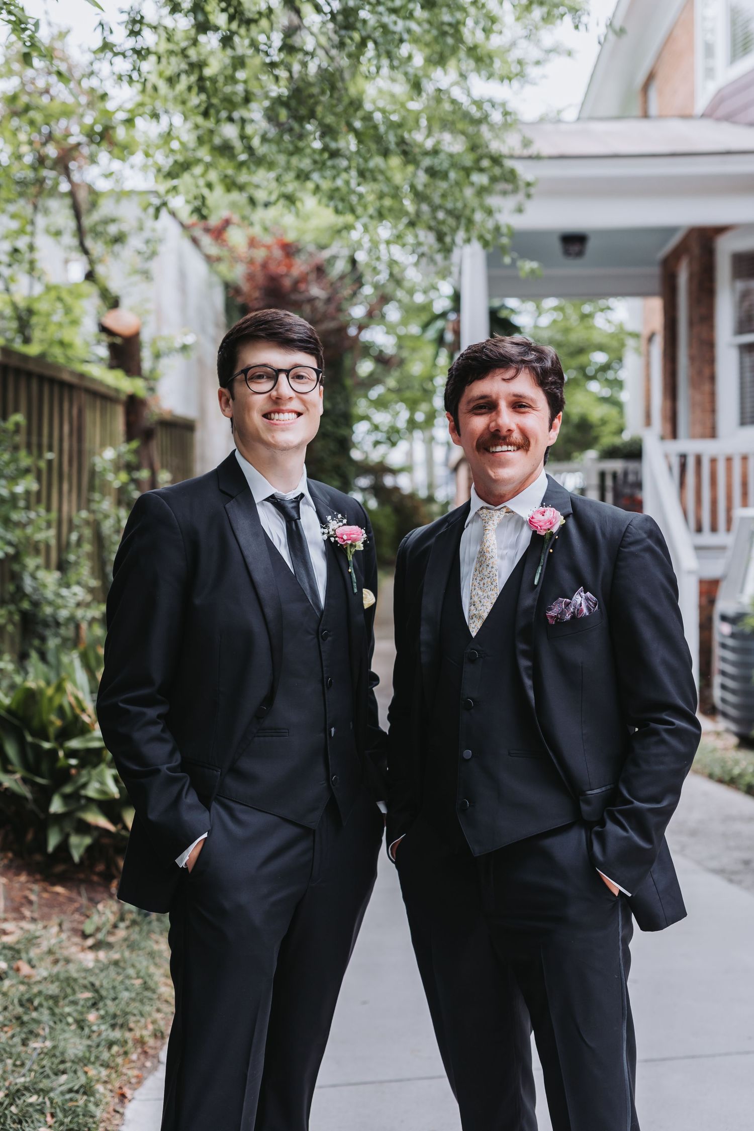 Two groomsmen in black suits with pink boutonnieres smile together outdoors on wedding day.