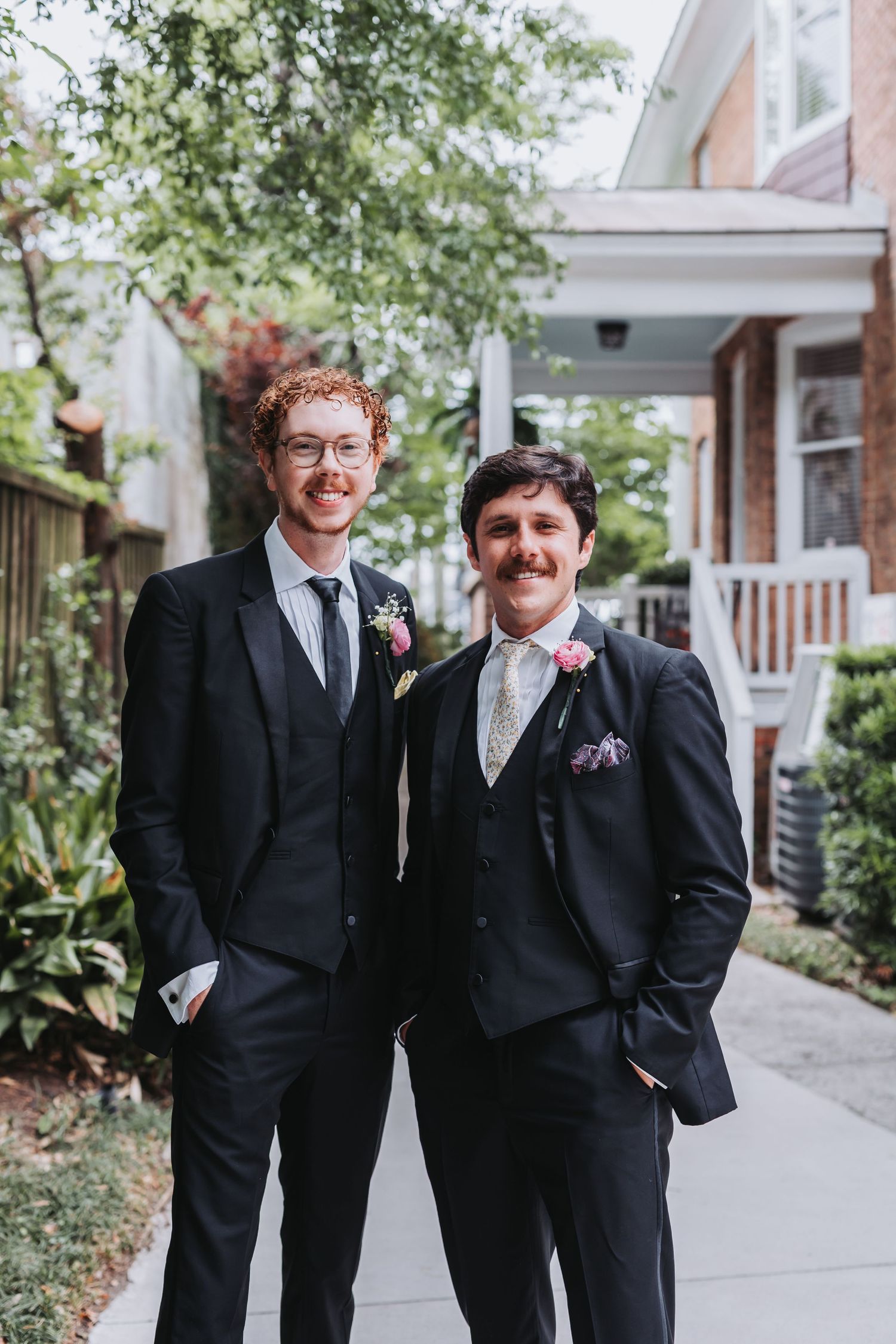 Two groomsmen in black suits with floral boutonnieres smile together outside before the wedding.