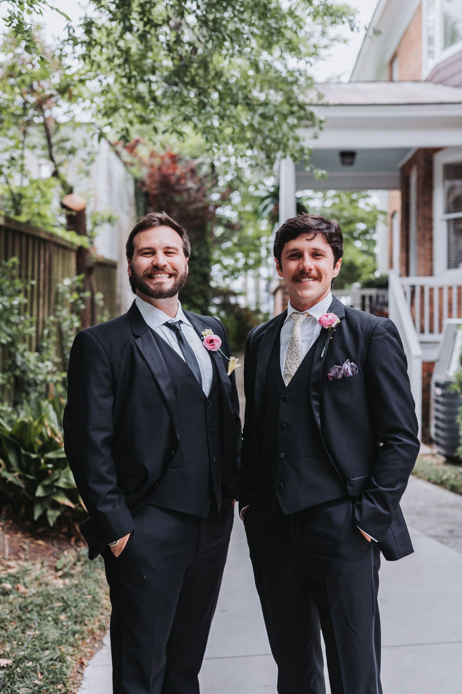 Two grooms in dark suits with boutonnieres smile together outdoors on their wedding day.