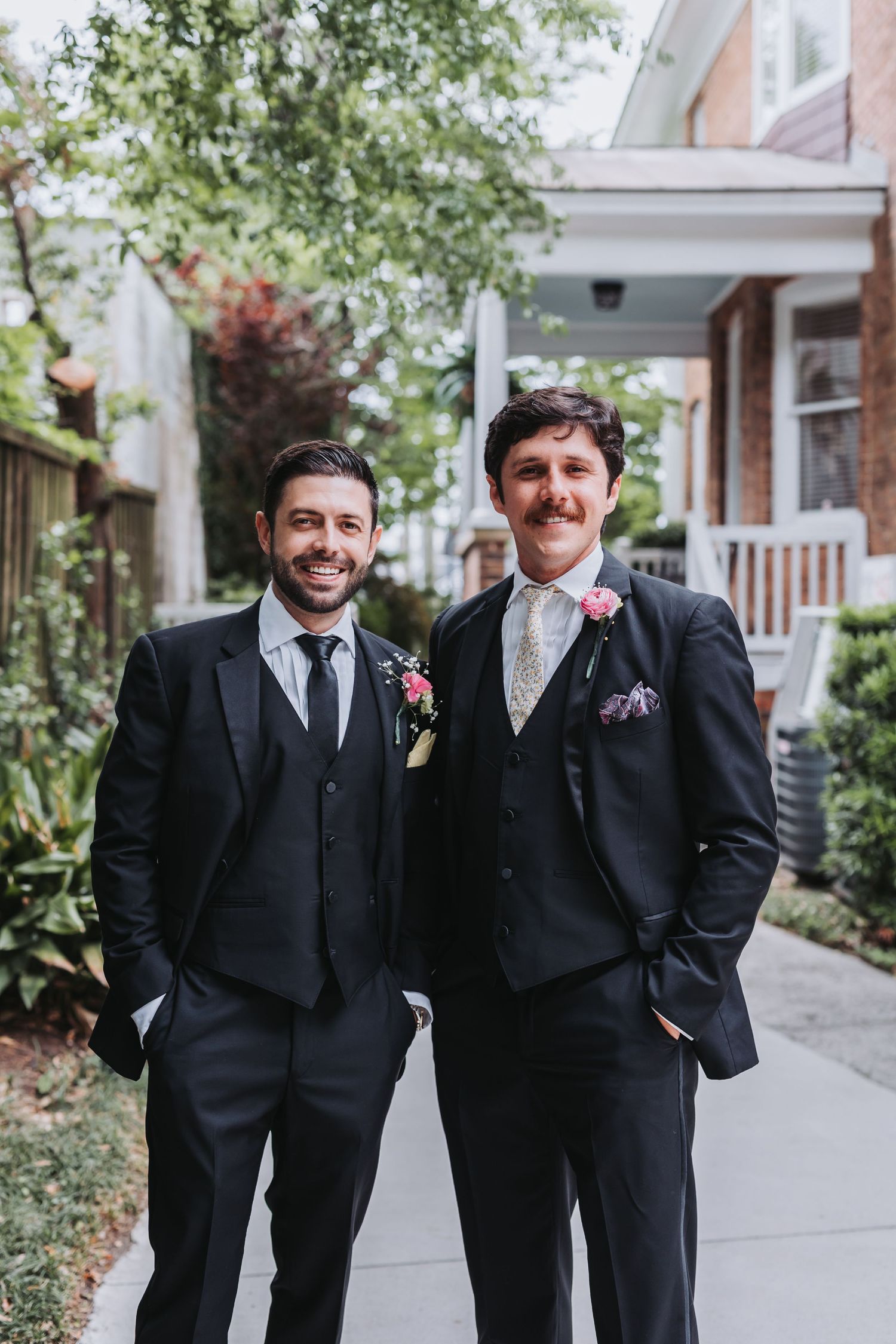 Two smiling groomsmen in black suits with floral boutonnieres pose together on a sunny sidewalk.
