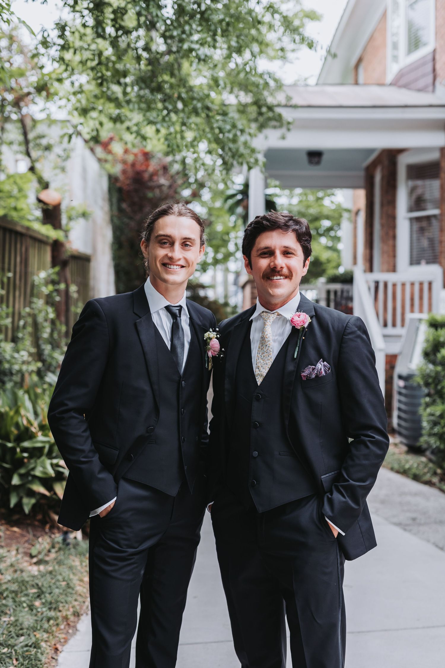 Two groomsmen in black suits with boutonnieres smile outdoors before a wedding ceremony.