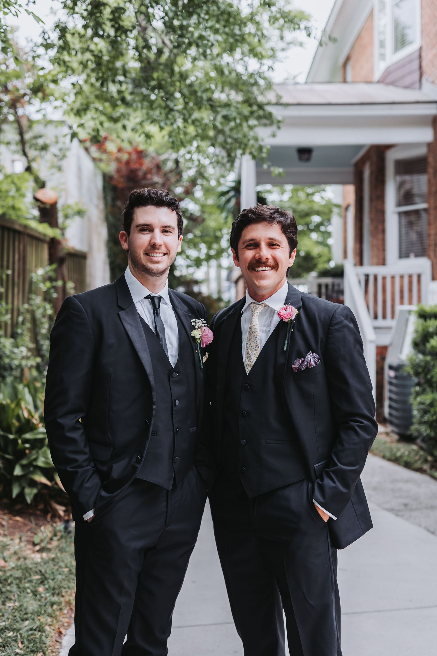 Two groomsmen in black suits with pink boutonnieres smiling outdoors before wedding ceremony.
