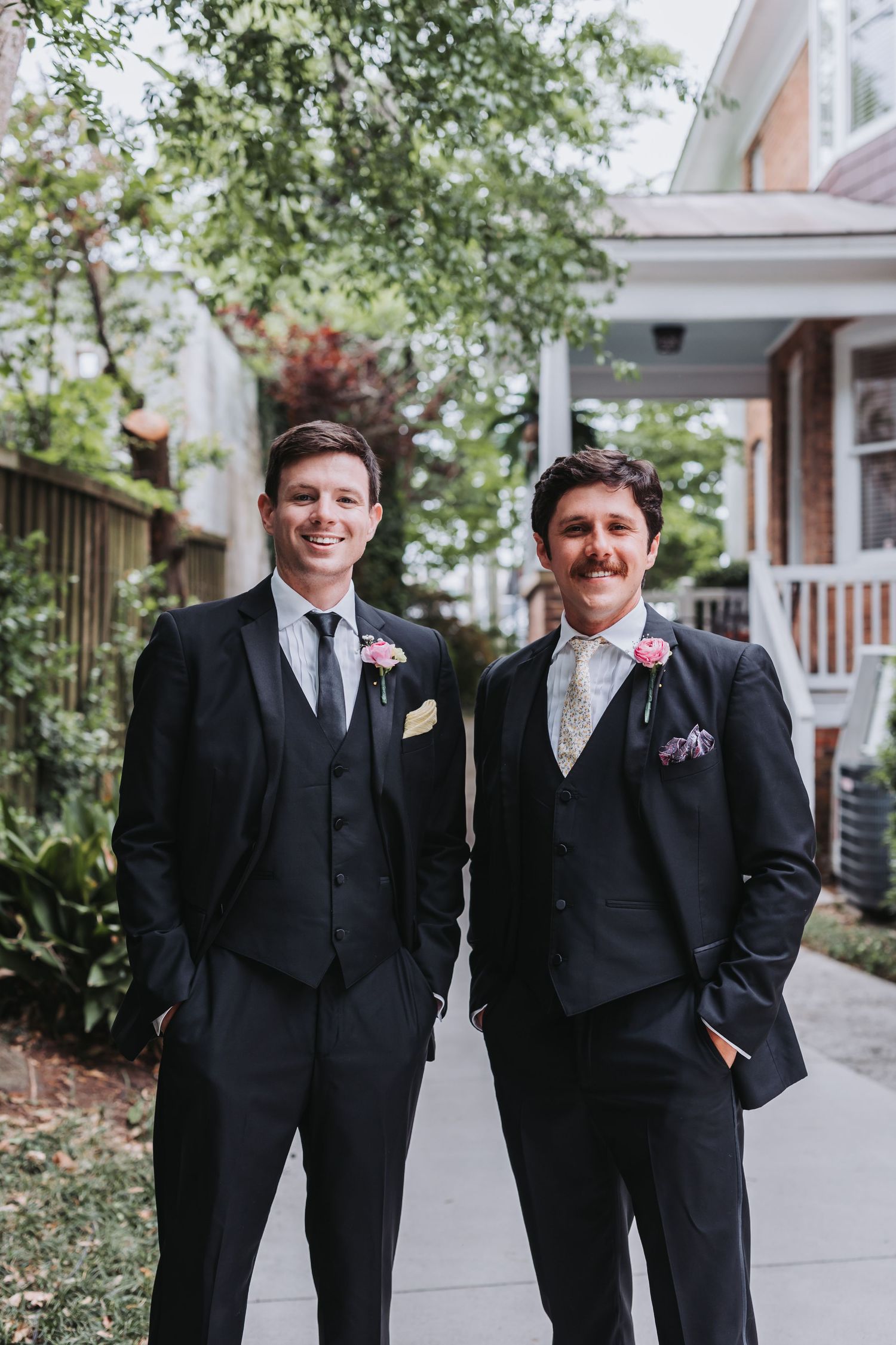 Two groomsmen in black suits with floral boutonnieres smiling outdoors at a wedding.