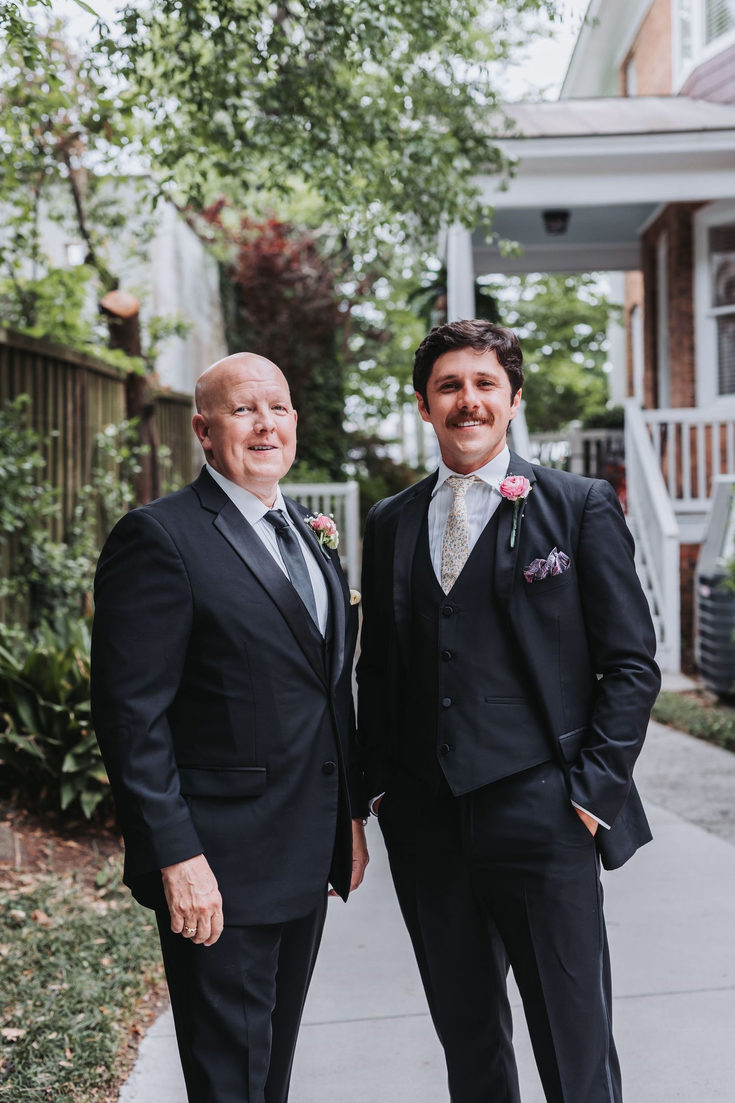 Groom and his father pose together in matching dark suits on the sidewalk before the wedding ceremony.