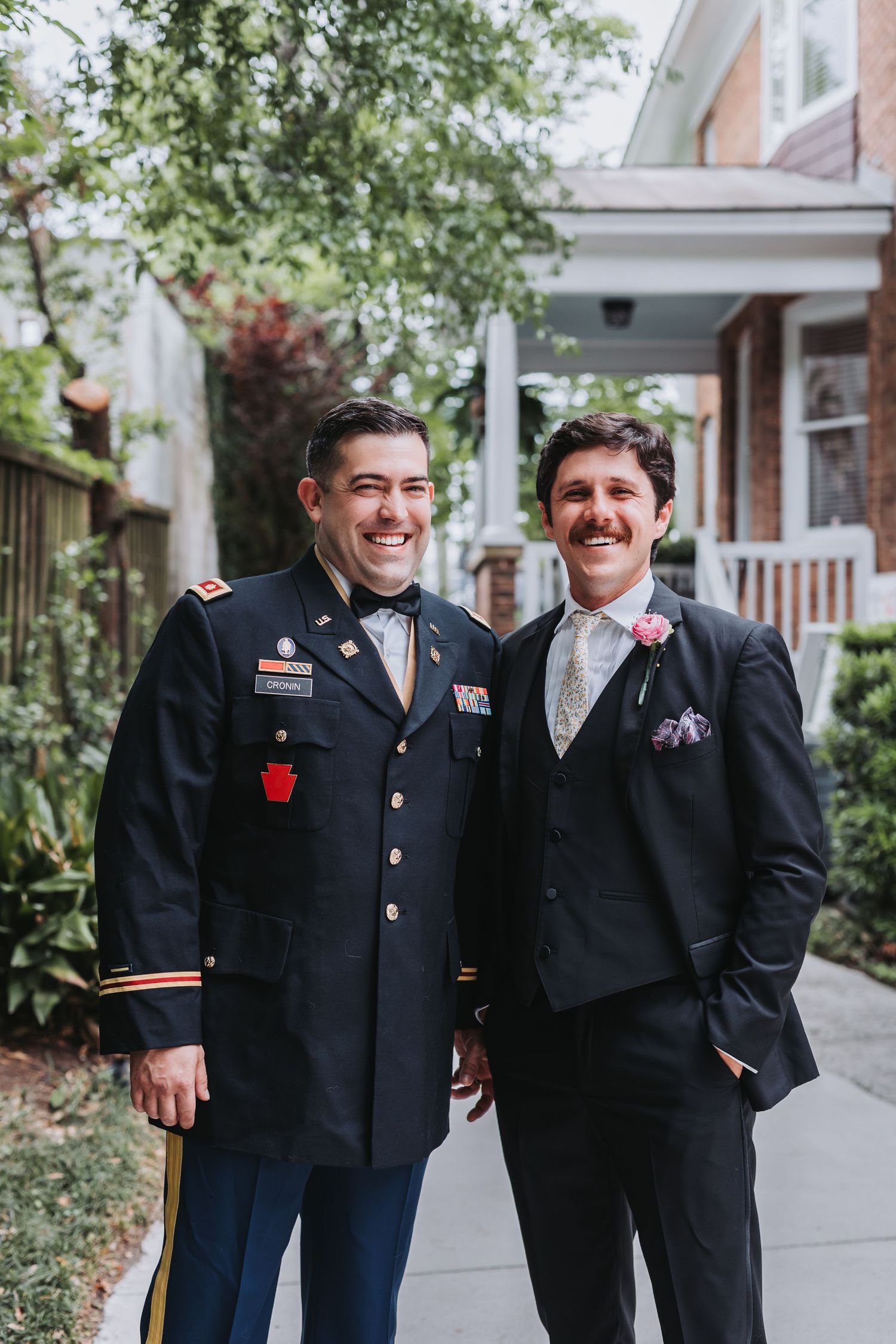 Two smiling men in formal attire, one in military dress uniform and one in a suit, posing together outdoors at a wedding.