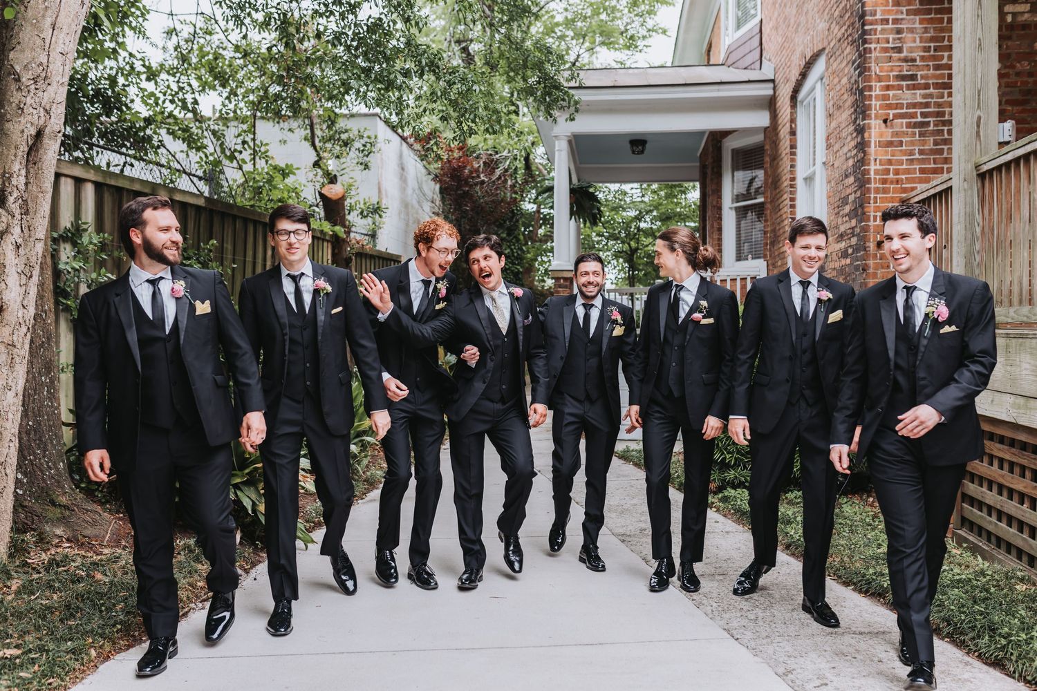 Stylish groom and groomsmen in matching black suits laugh and walk together outdoors at wedding.