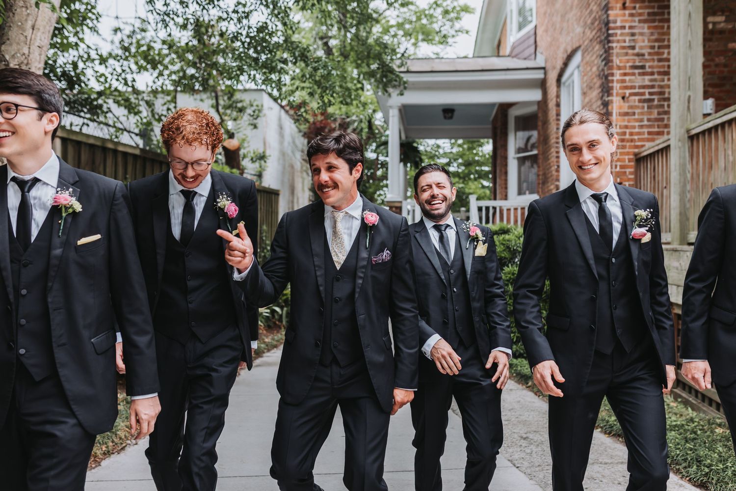 Groomsmen in matching black suits with boutonnieres walk and laugh together outdoors before a wedding ceremony.