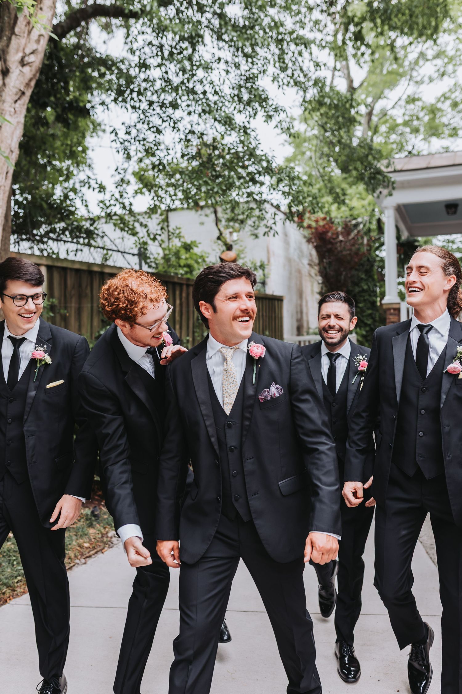 Groom and groomsmen laughing and walking together in black suits with floral boutonnieres at outdoor wedding.