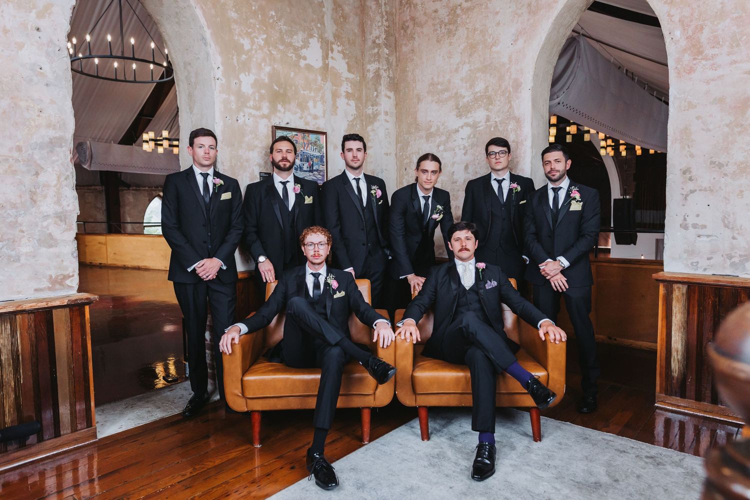 Groom and groomsmen pose in black suits on leather sofa in elegant rustic wedding venue.