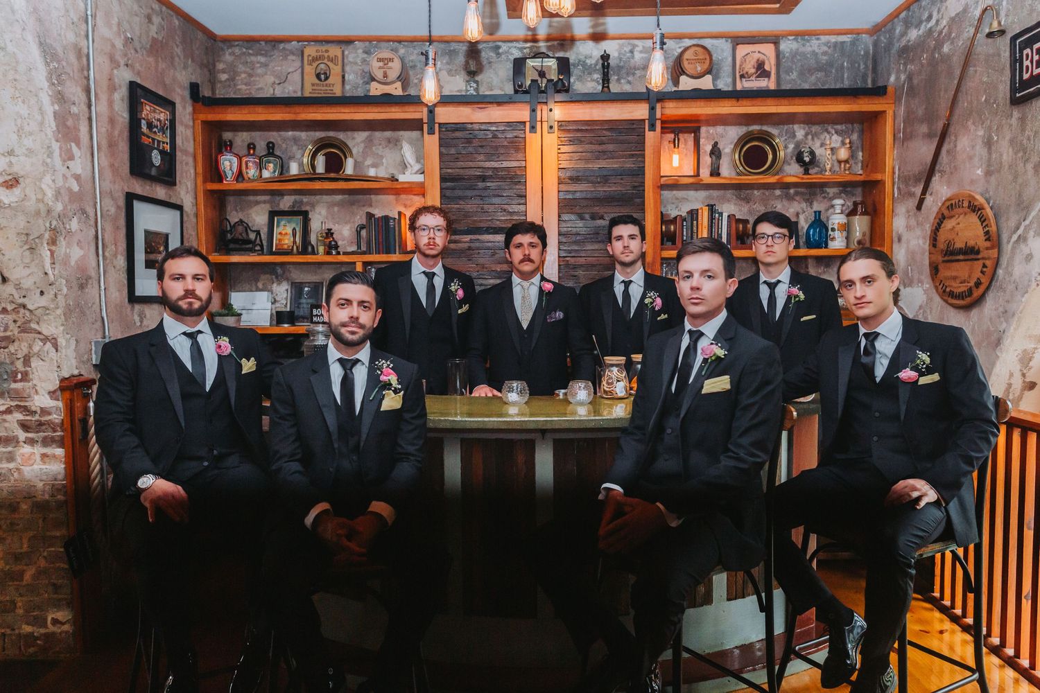 Seven groomsmen in black suits pose together at a rustic bar venue for wedding photo.