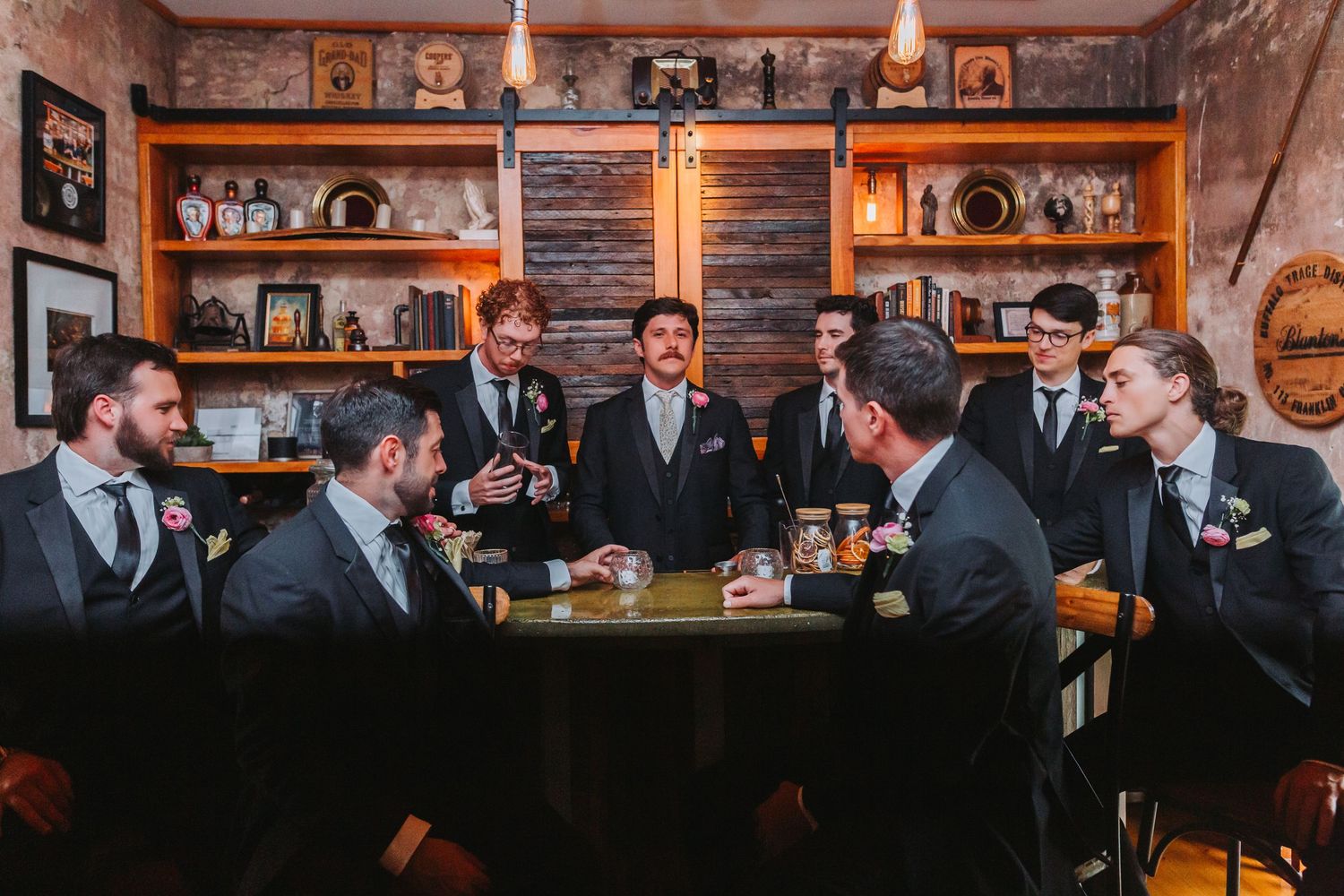 Groom addresses groomsmen in dark suits gathered around a table in a warmly lit rustic room.