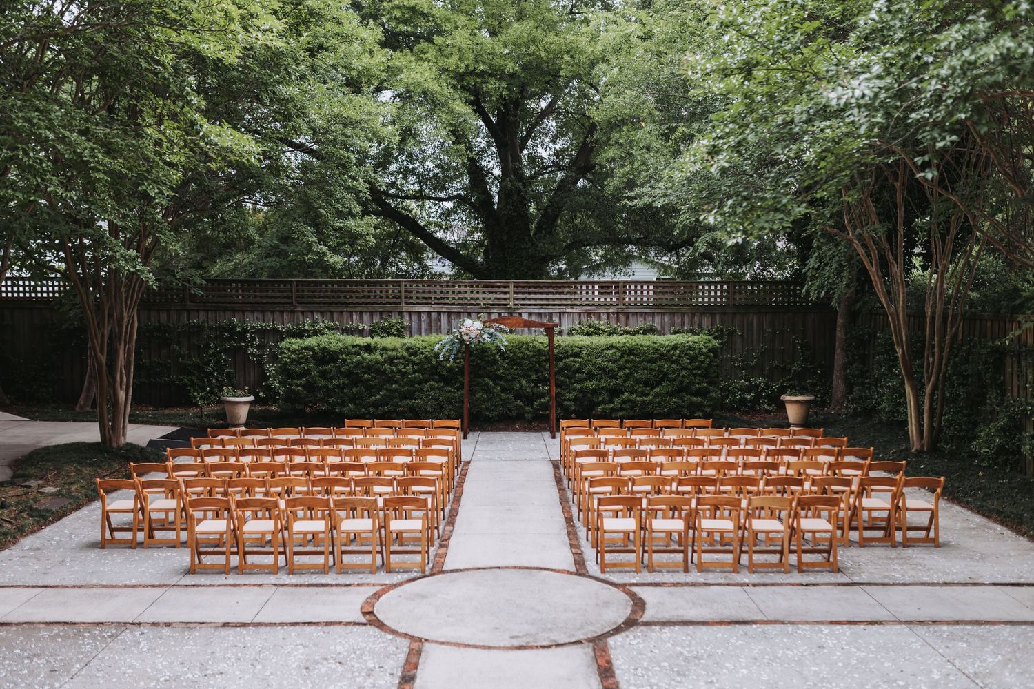 Elegant outdoor wedding ceremony venue with wooden folding chairs arranged under lush green tree canopy.