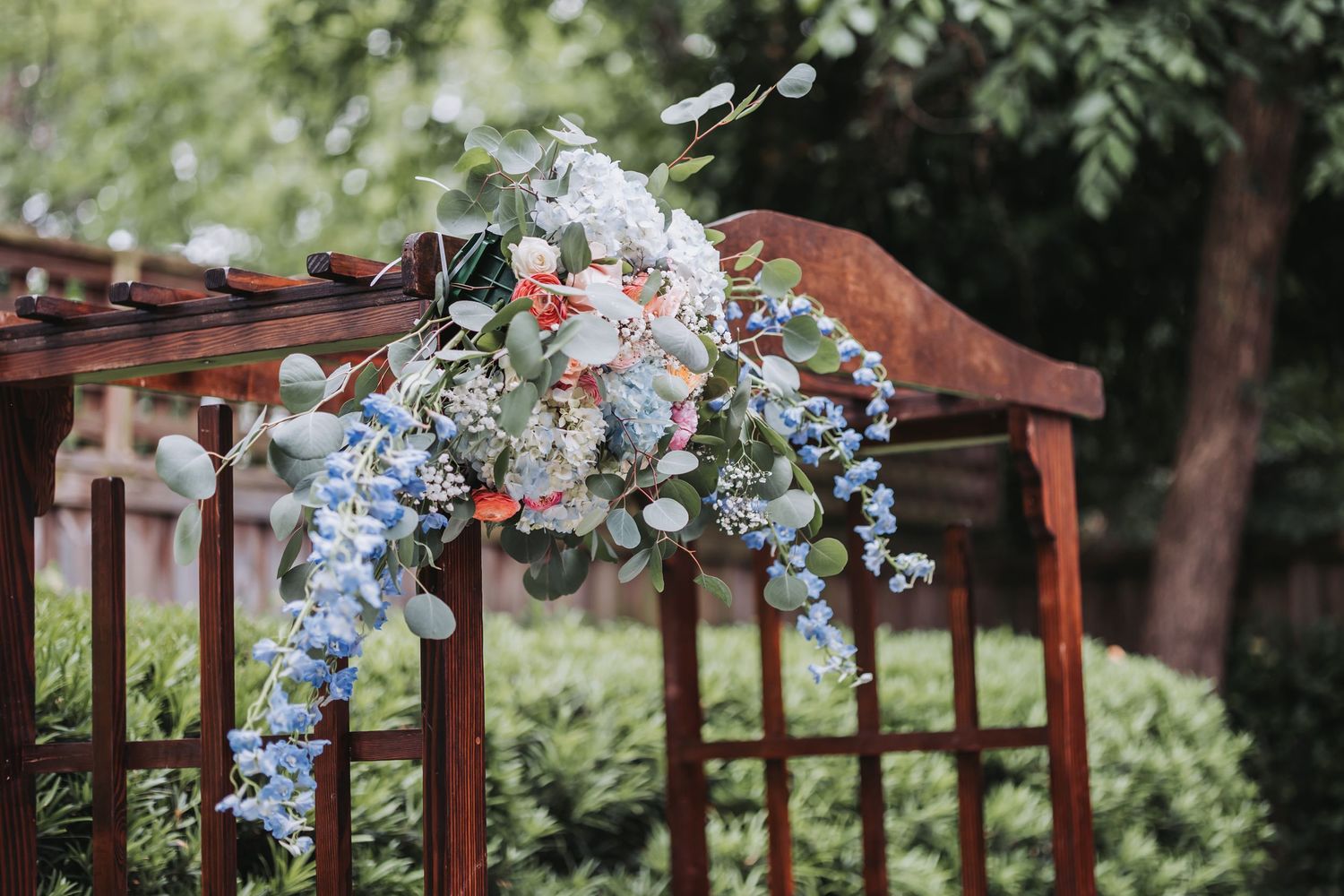 Elegant blue and white floral arrangement with delphinium and eucalyptus draped over wooden wedding arch.