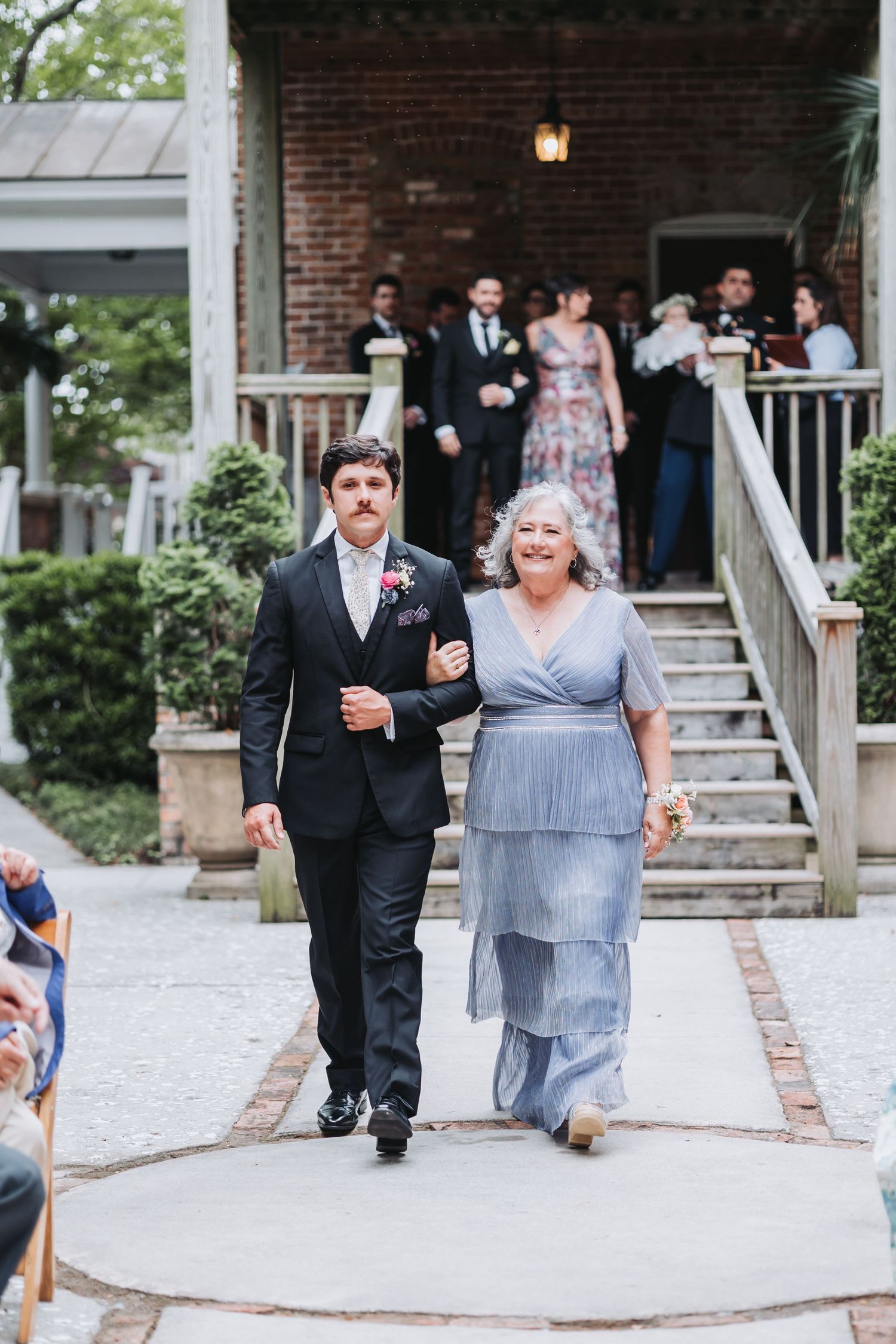 Groom in black suit escorts older woman in blue tiered dress down wedding ceremony aisle.