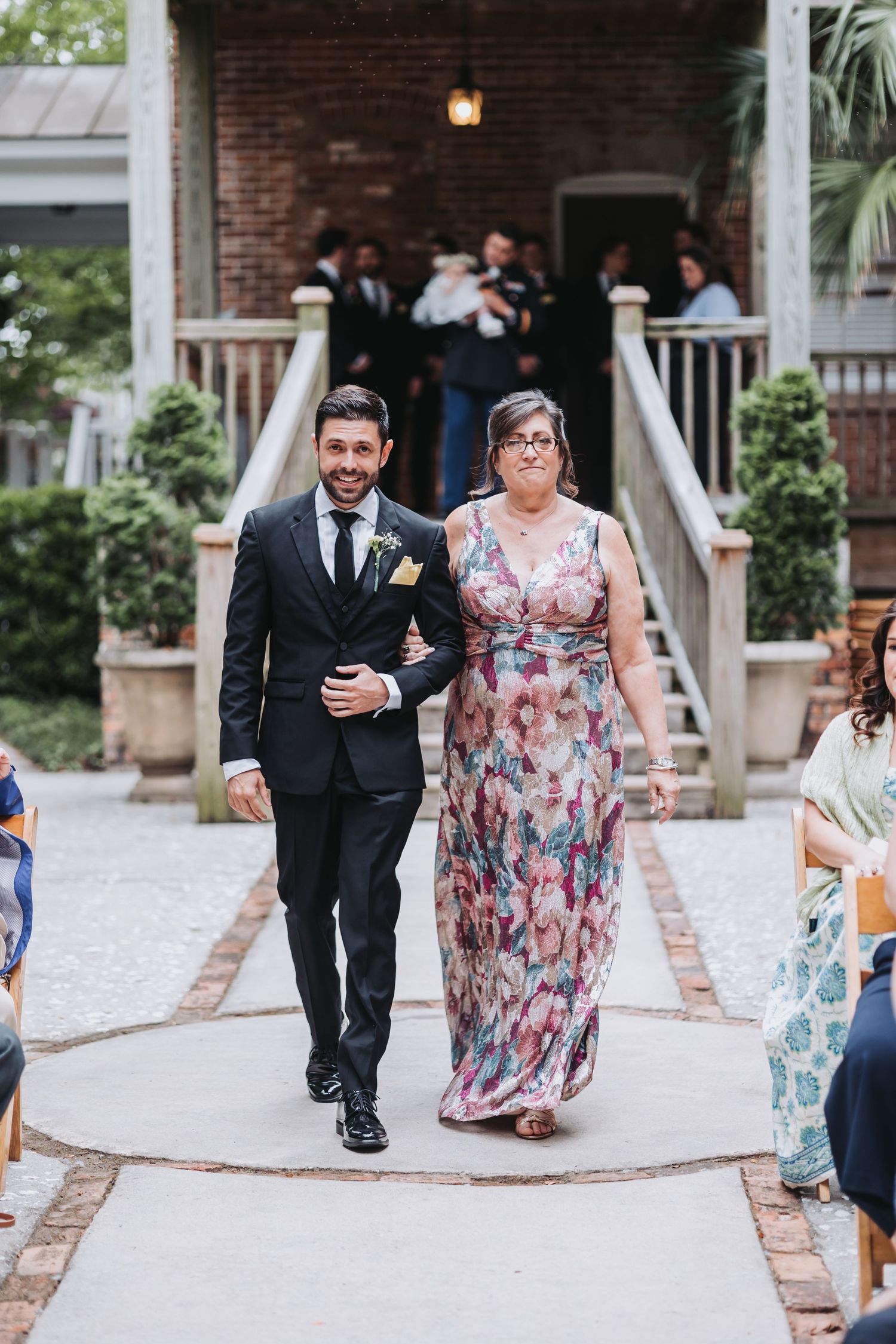 Groom in black suit escorts woman in floral dress down wedding ceremony aisle at outdoor venue.