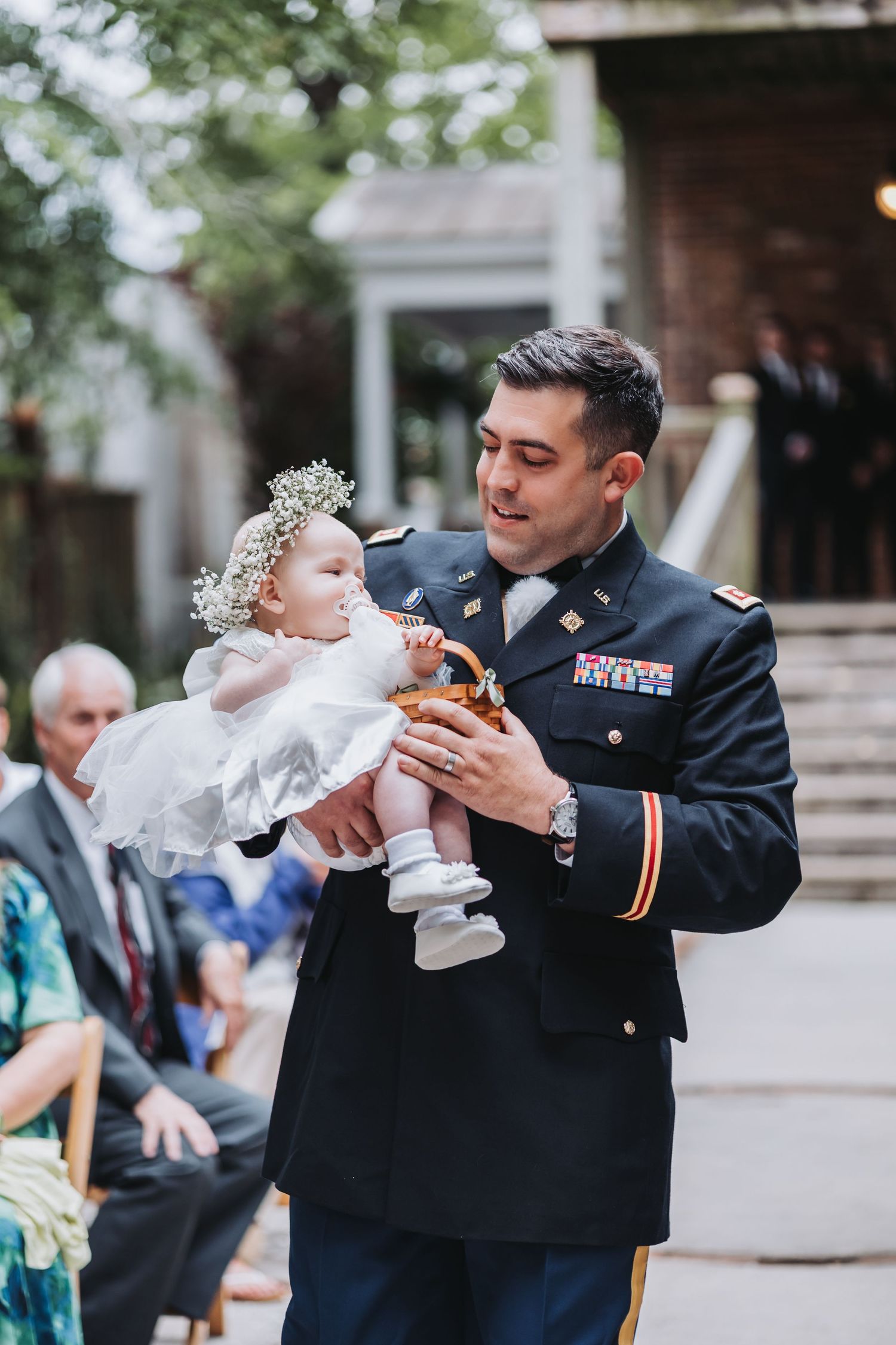Military officer in dress uniform carries flower girl down wedding ceremony aisle.
