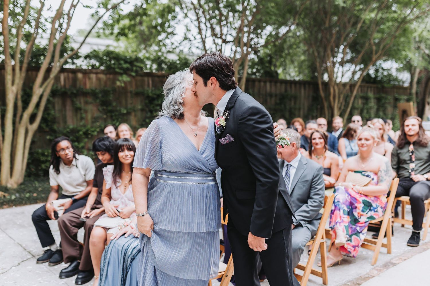 Groom in dark suit shares tender kiss with mother seated in chair during outdoor wedding ceremony.