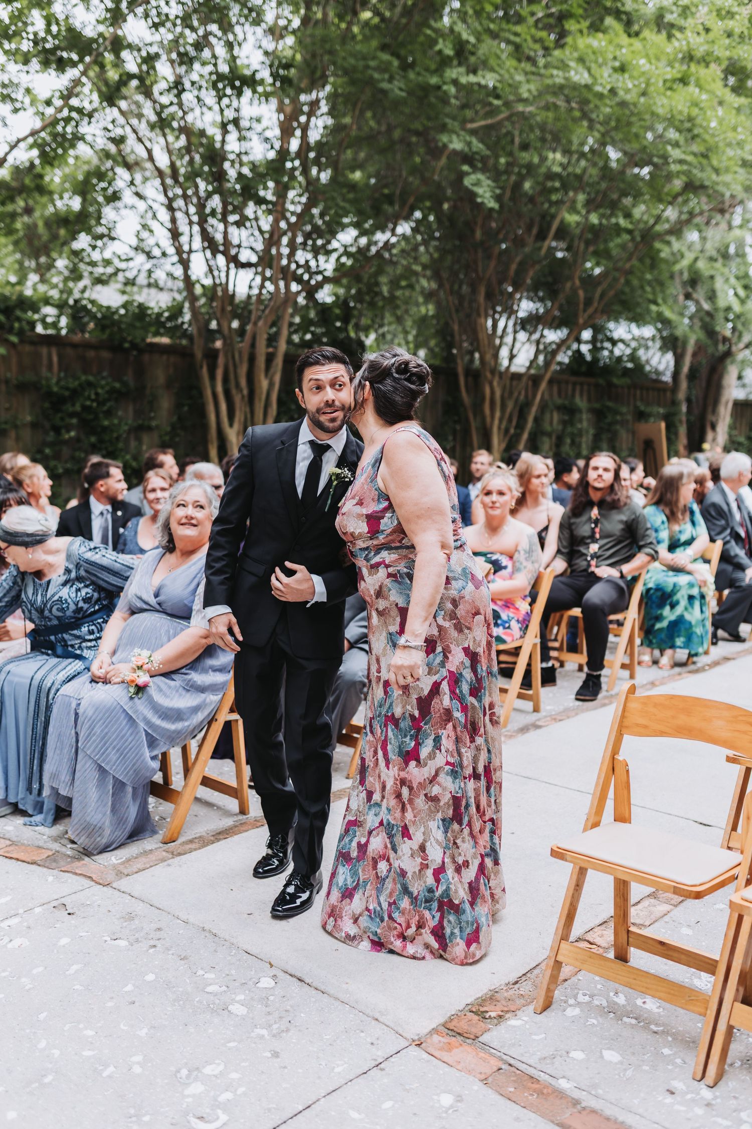 A woman in a floral gown kisses a man in a black suit on the cheek during an outdoor wedding ceremony.