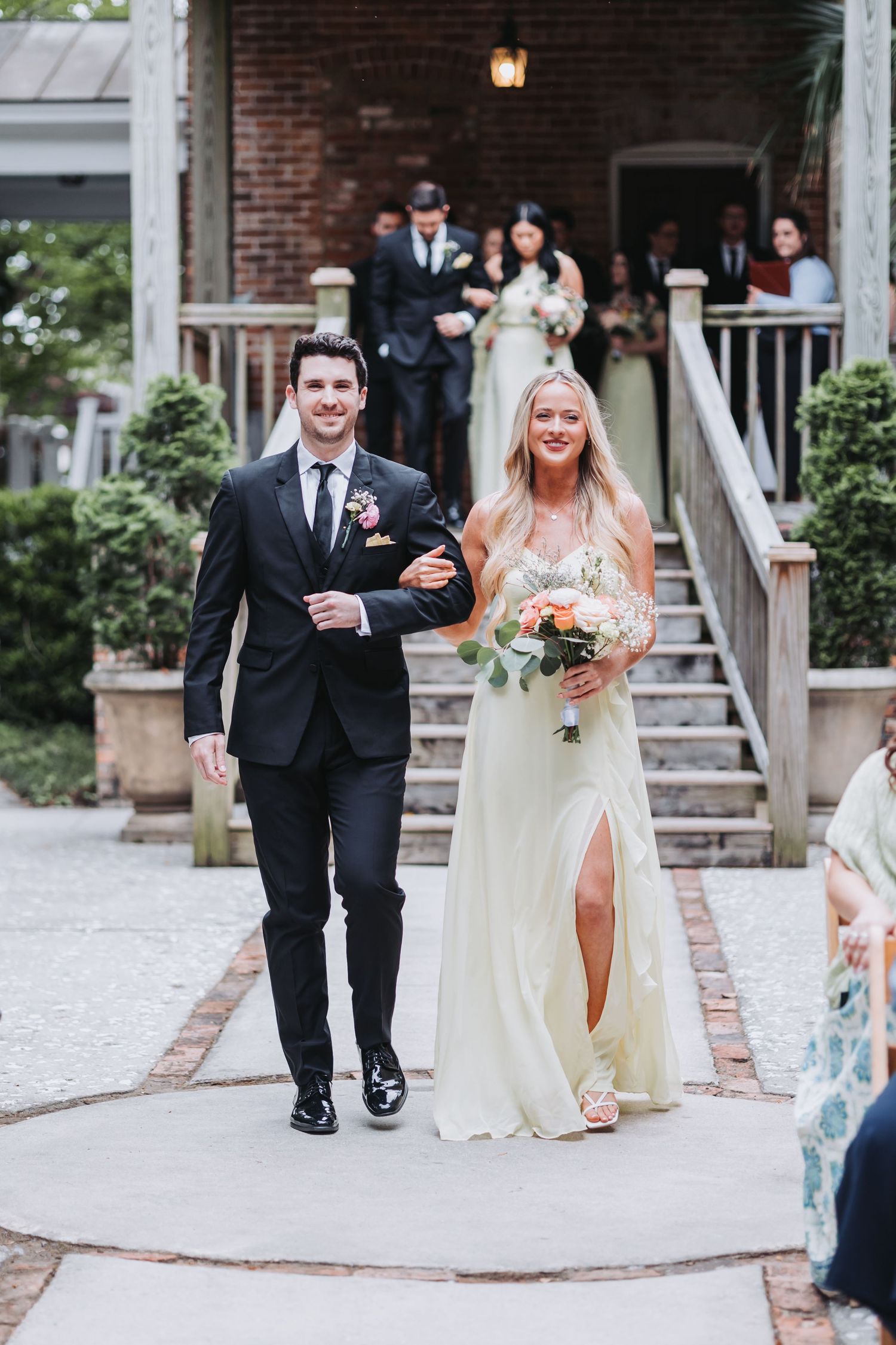 A smiling bridesmaid in a white gown walks down the aisle with a groomsman in a black suit at an outdoor wedding.