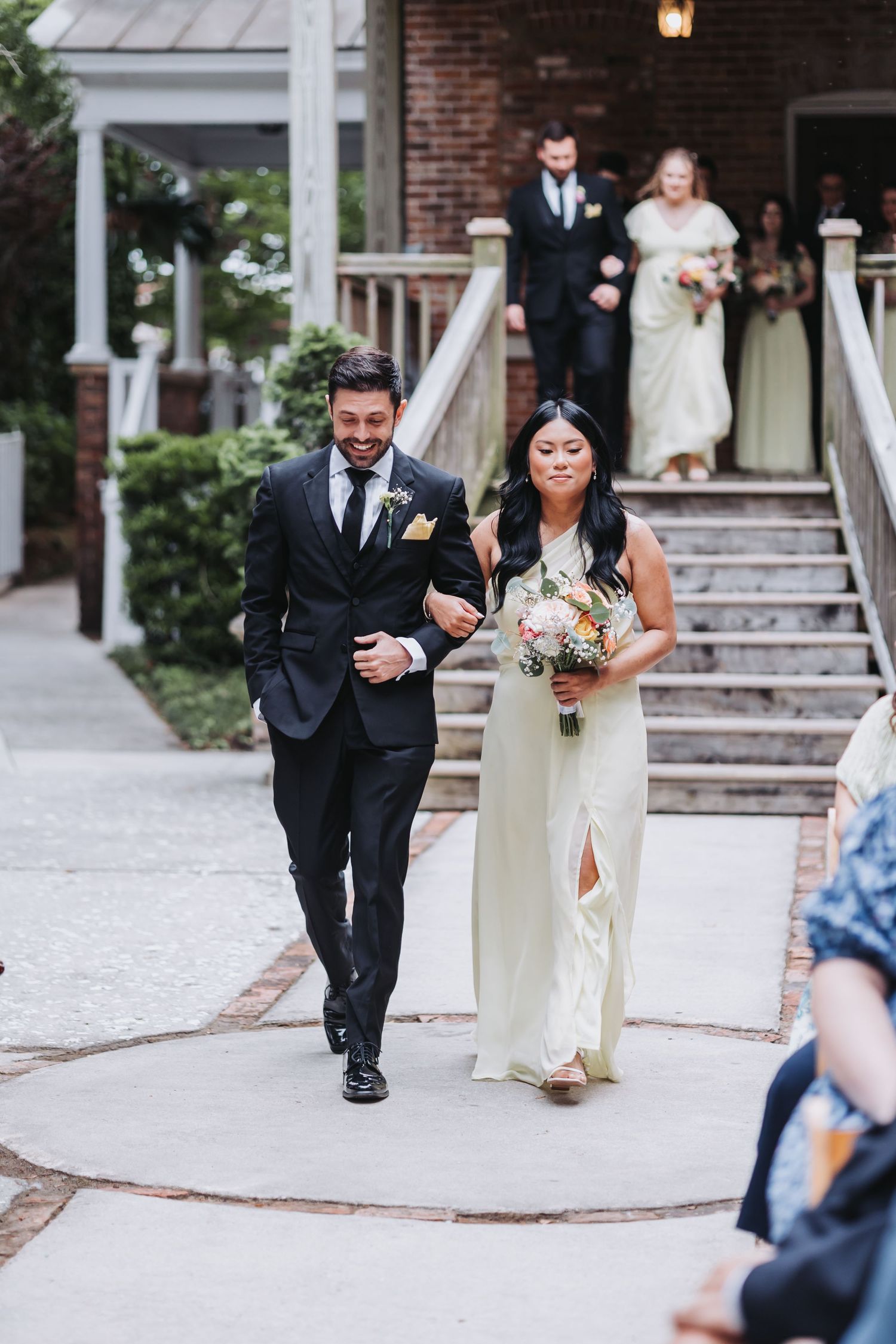 Groomsman in black suit escorts bridesmaid in sage green gown down outdoor wedding aisle.