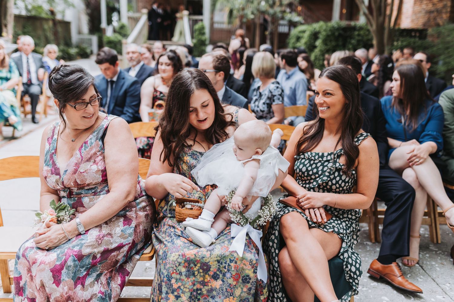 Three women sit together at a wedding ceremony, one holding and smiling at a baby dressed in white.