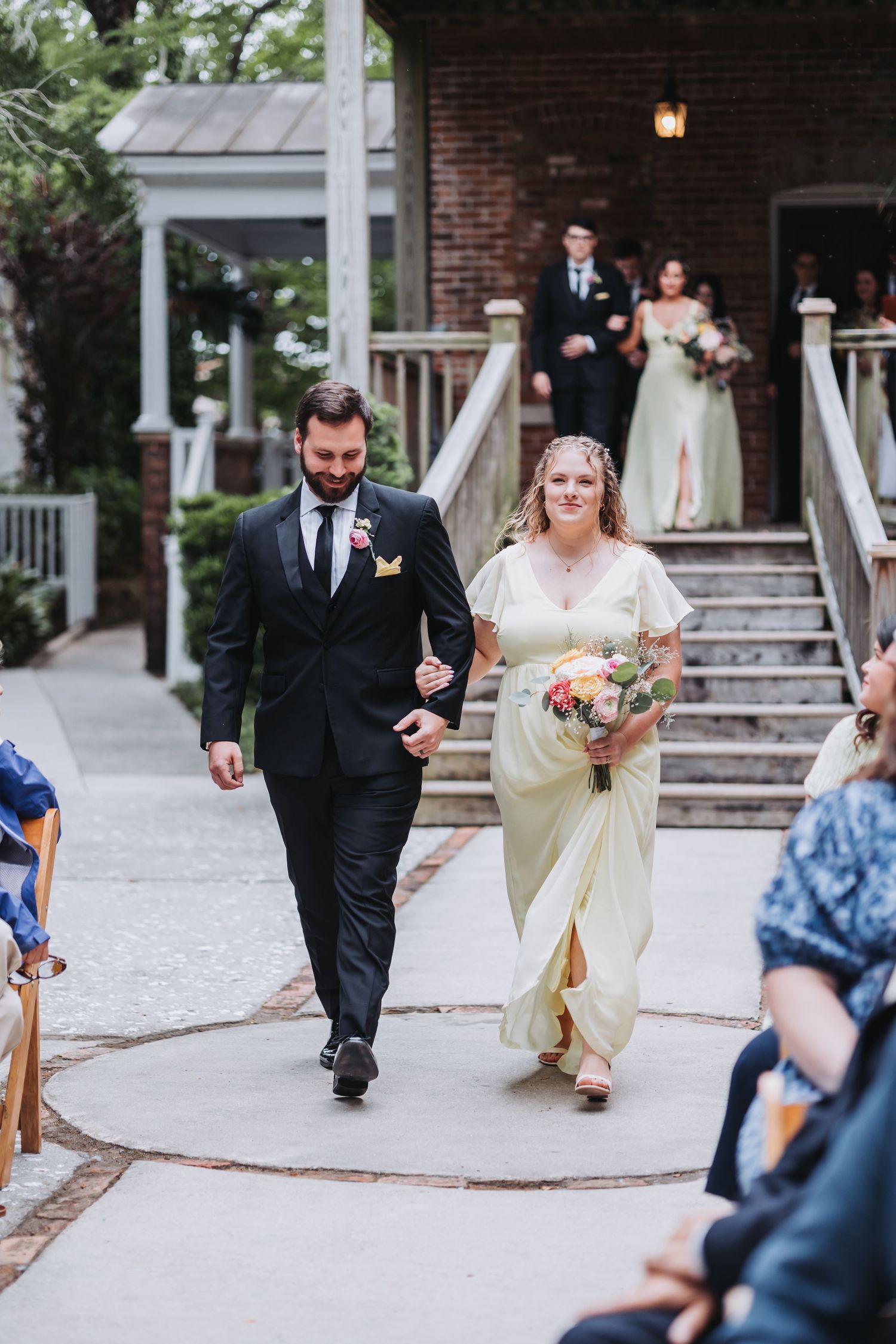 A bridesmaid in a light yellow gown walks arm-in-arm with a bearded groomsman down an outdoor wedding aisle.
