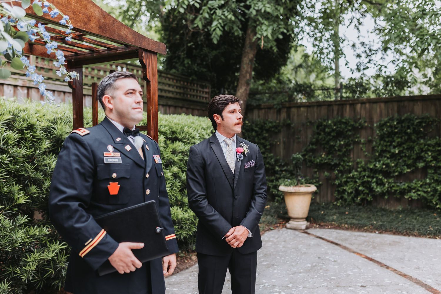 A military officer in uniform stands beside a groom in a gray suit waiting at an outdoor wedding ceremony.