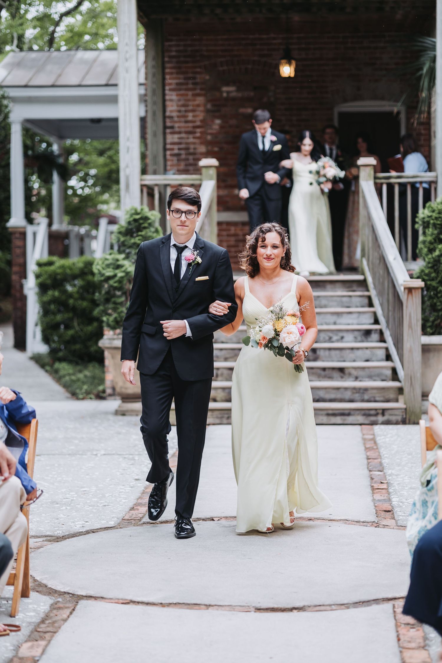 Groomsman in black suit walks bridesmaid in sage dress down outdoor wedding ceremony aisle.