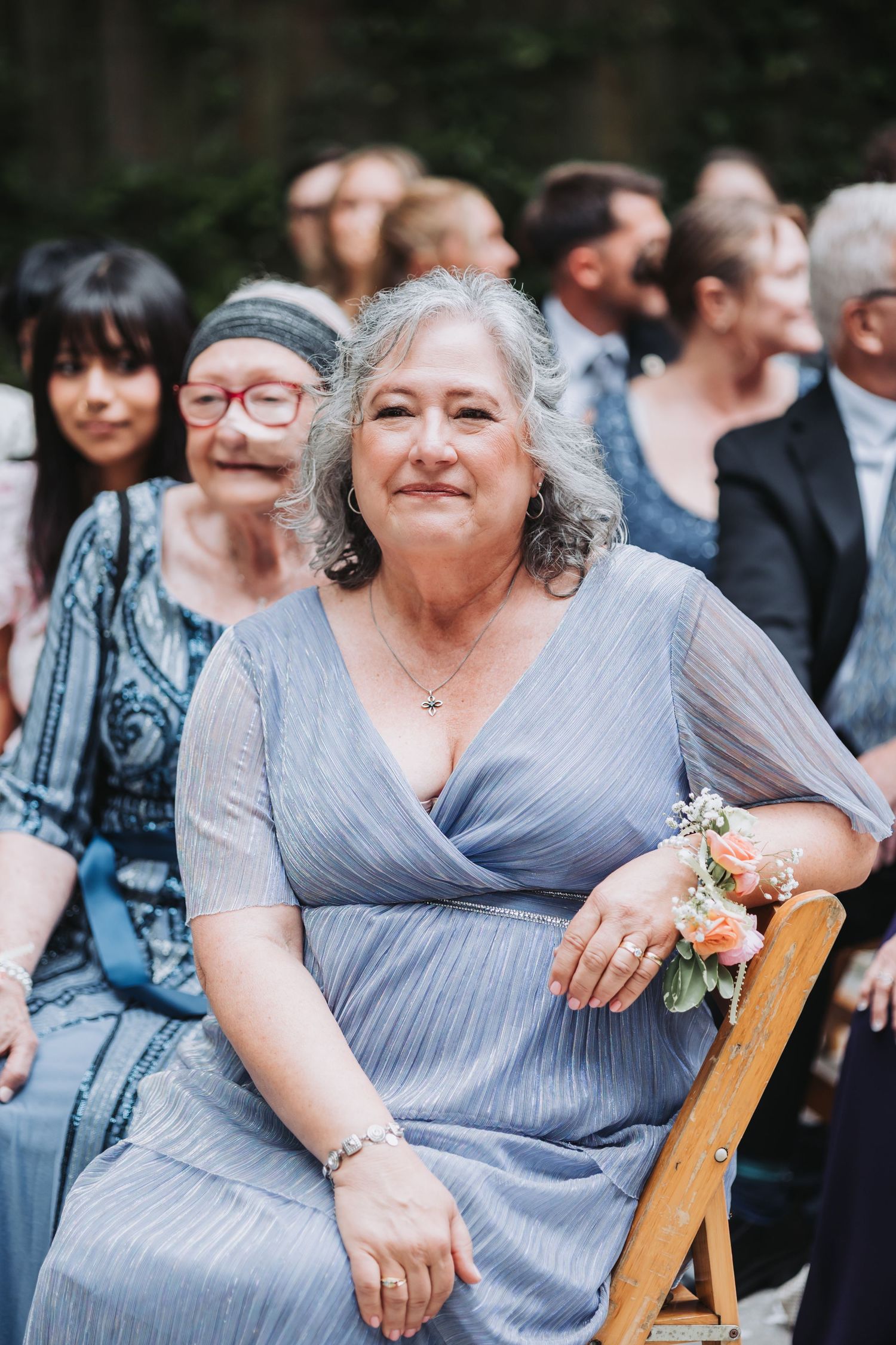 Elegant older woman in blue dress with floral corsage smiling at outdoor wedding ceremony.