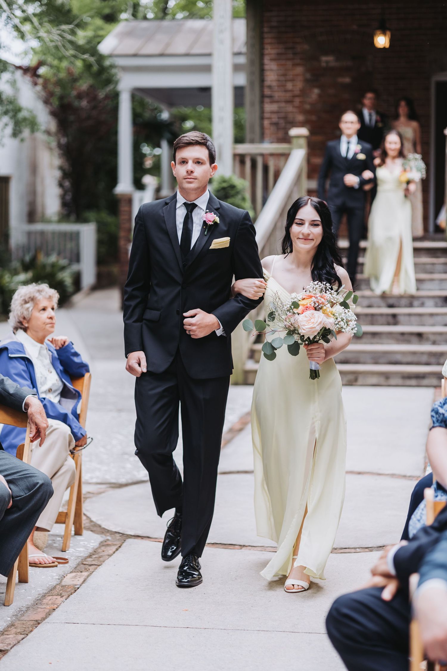 Groomsman in black suit walks bridesmaid in sage green dress holding bouquet down the aisle.