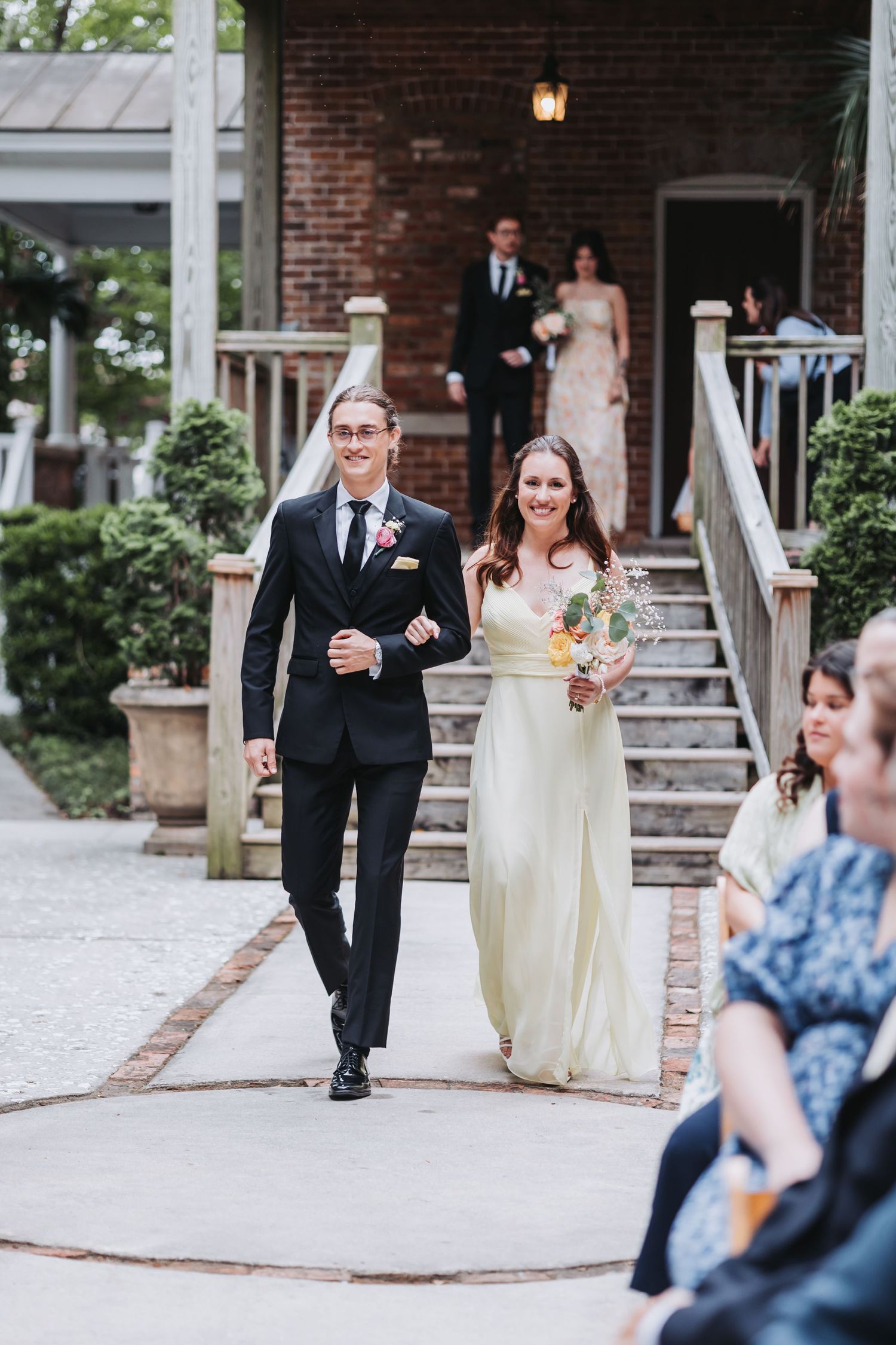A smiling bridesmaid in a pale yellow gown walks arm-in-arm with a tall groomsman down an outdoor wedding aisle.