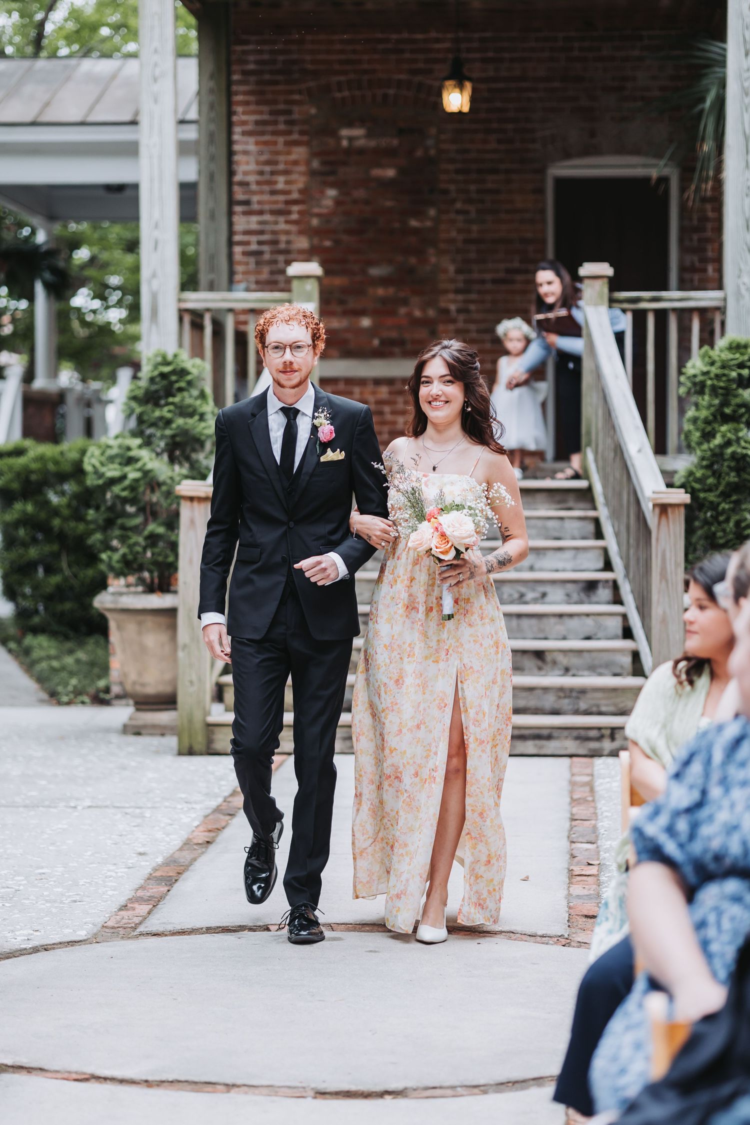 Groomsman escorts bridesmaid in floral dress down outdoor aisle at garden wedding ceremony.
