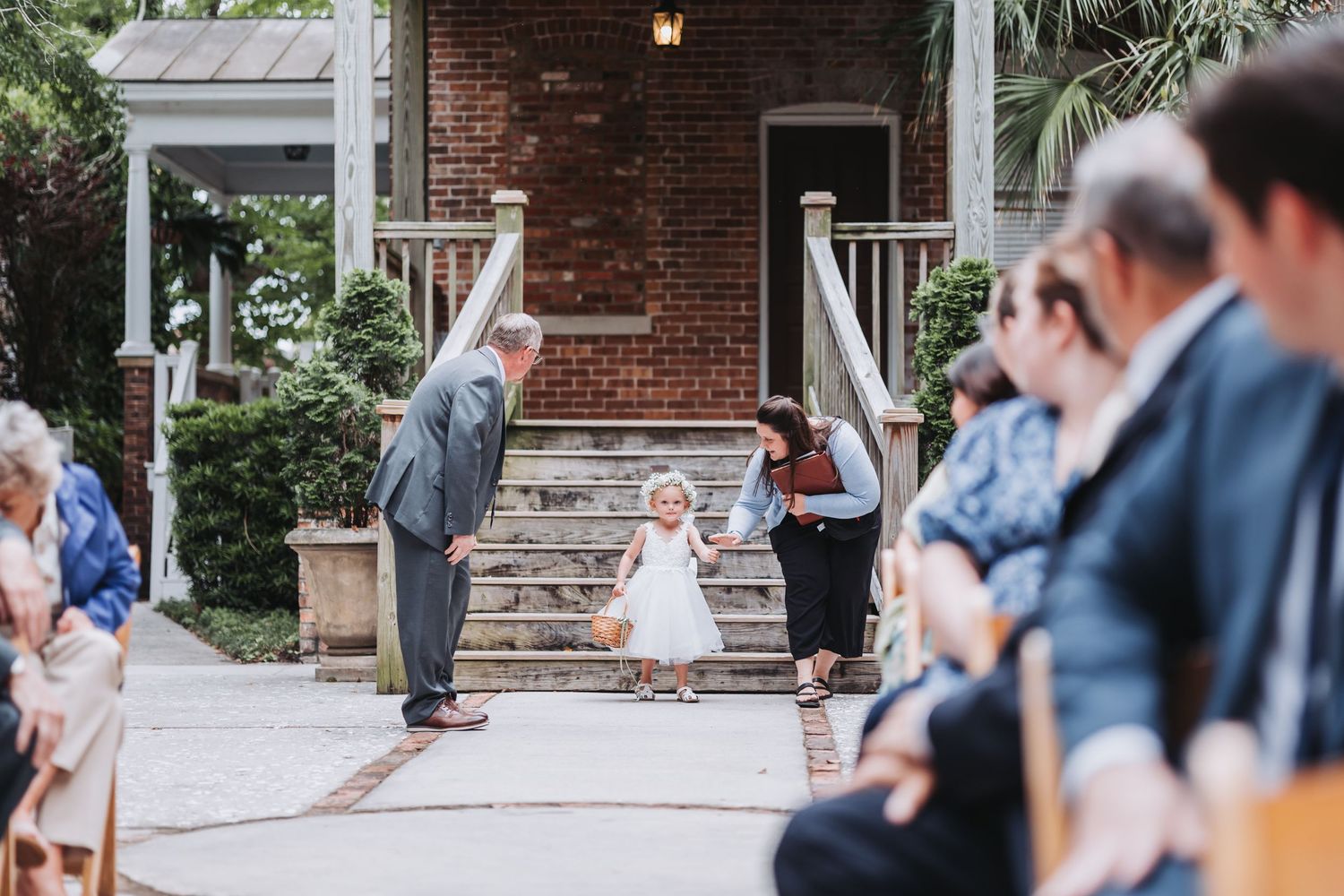 Adorable flower girl in white dress walks down aisle at outdoor wedding ceremony.
