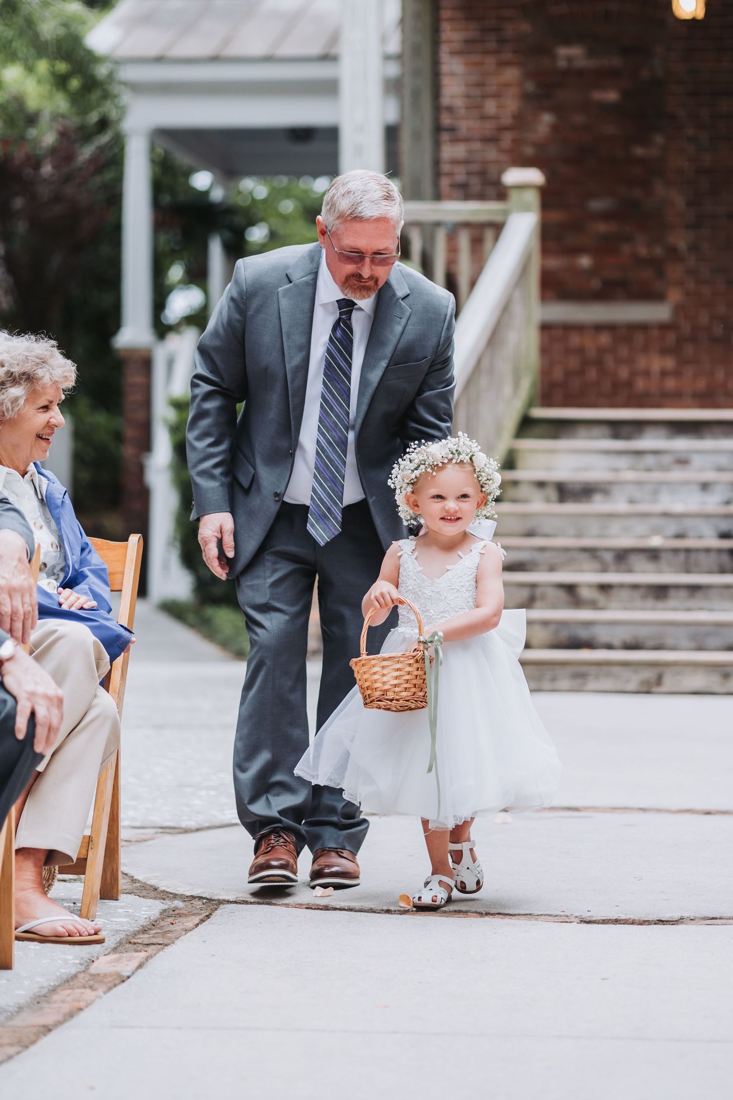 Elderly man escorts young flower girl in white dress down wedding aisle carrying wicker basket.