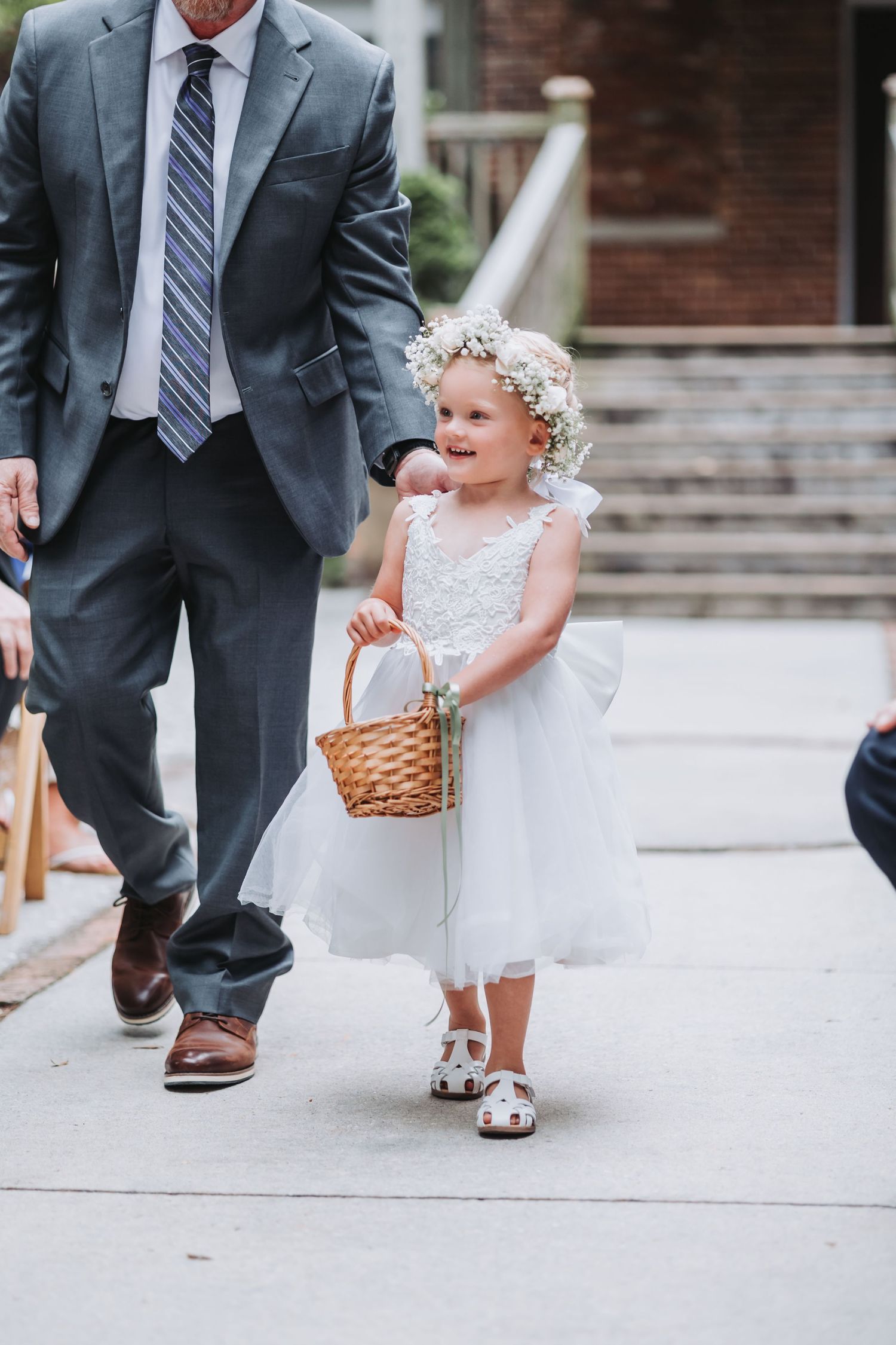 Adorable flower girl in white dress with floral crown carries basket down wedding aisle.