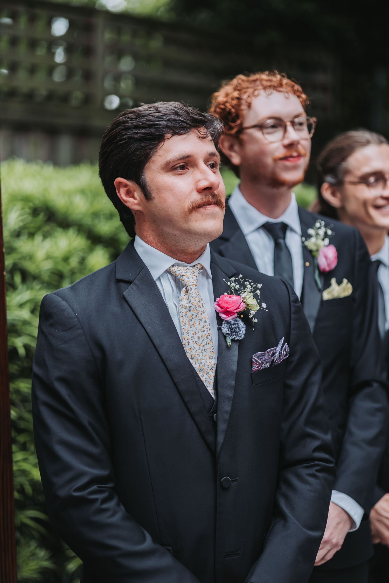 Handsome groom with mustache in dark suit watches bride walk down the aisle.