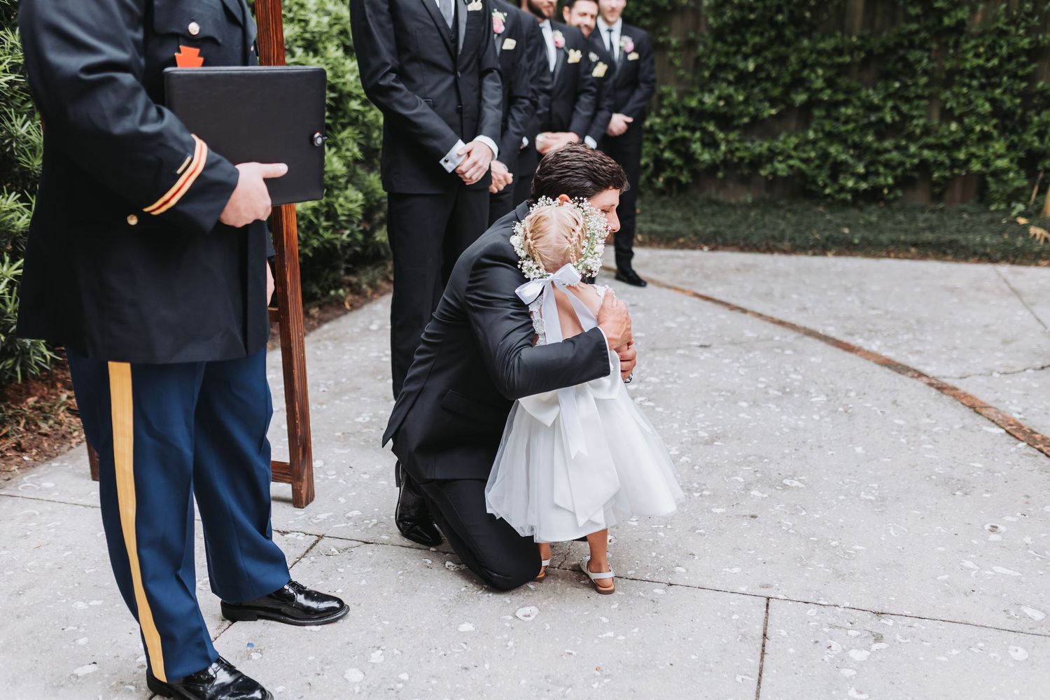 Groom kneels to embrace a young flower girl outside during the wedding ceremony while groomsmen stand nearby.