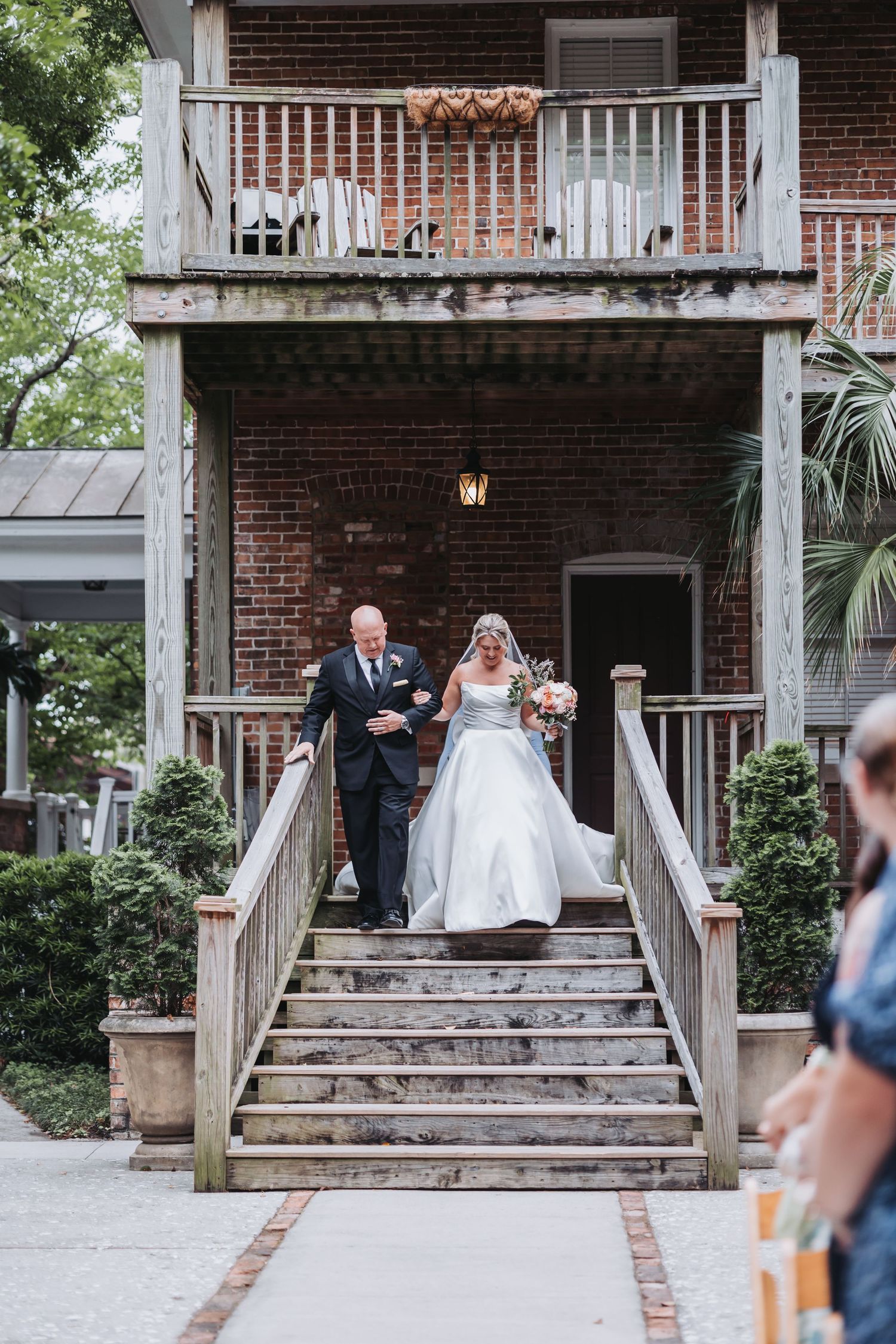 Bride in white gown descends brick building steps with escort holding colorful bouquet at outdoor wedding.
