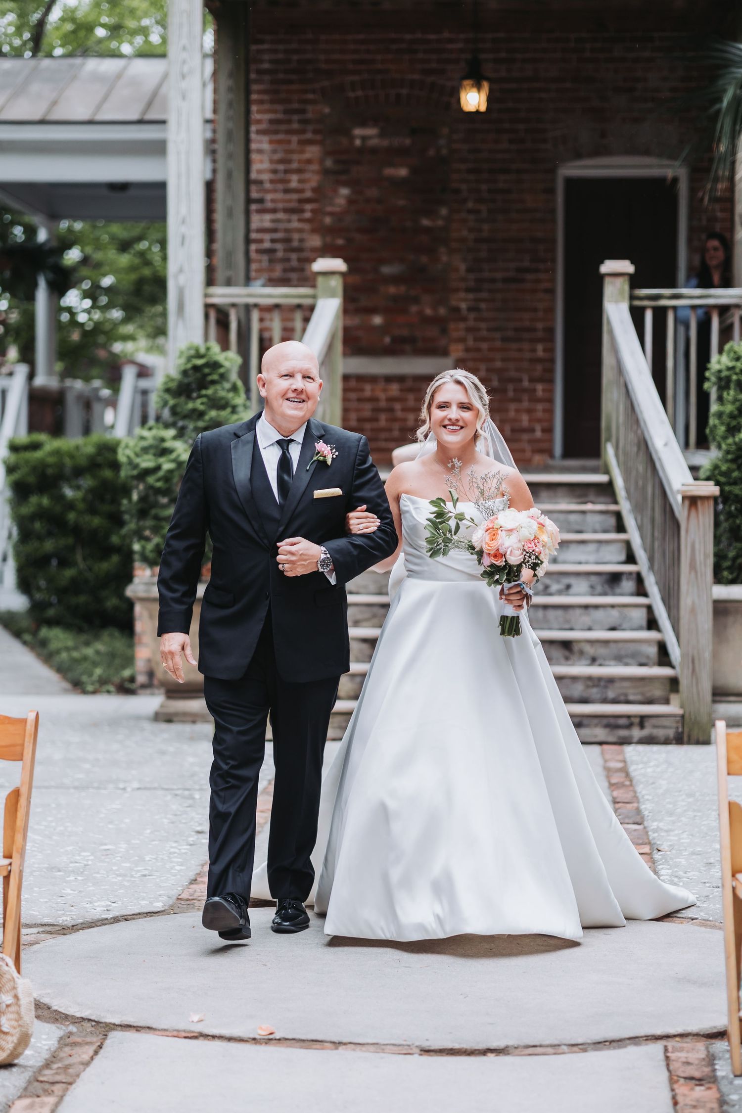 Bride in ball gown walks down outdoor aisle with escort at brick venue wedding.